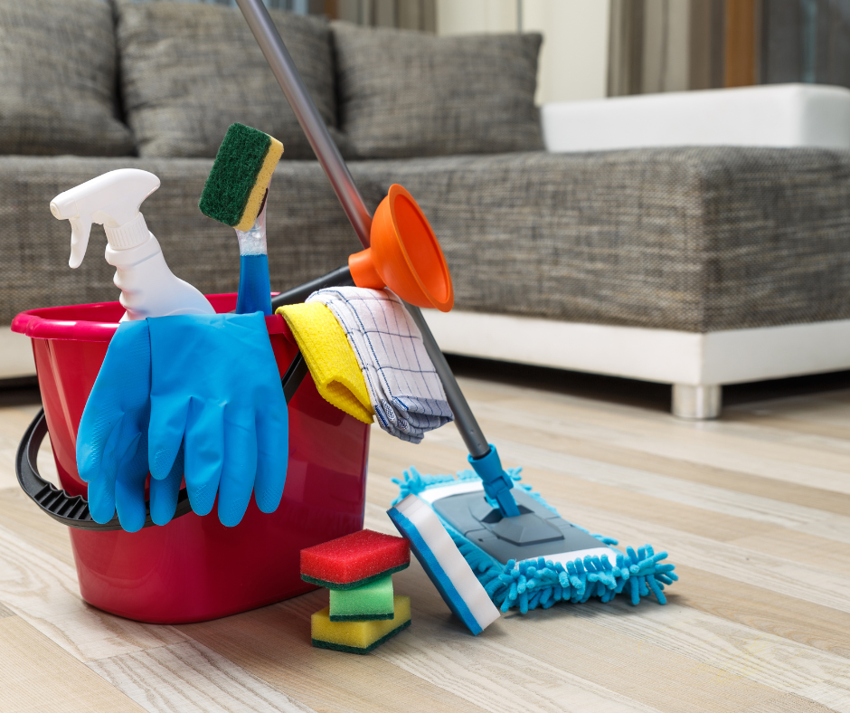 Cleaning supplies including a red bucket, spray bottle, rubber gloves, sponge, cloth, mop, and floor scrubber on a wooden floor in a living room with a gray sofa.
