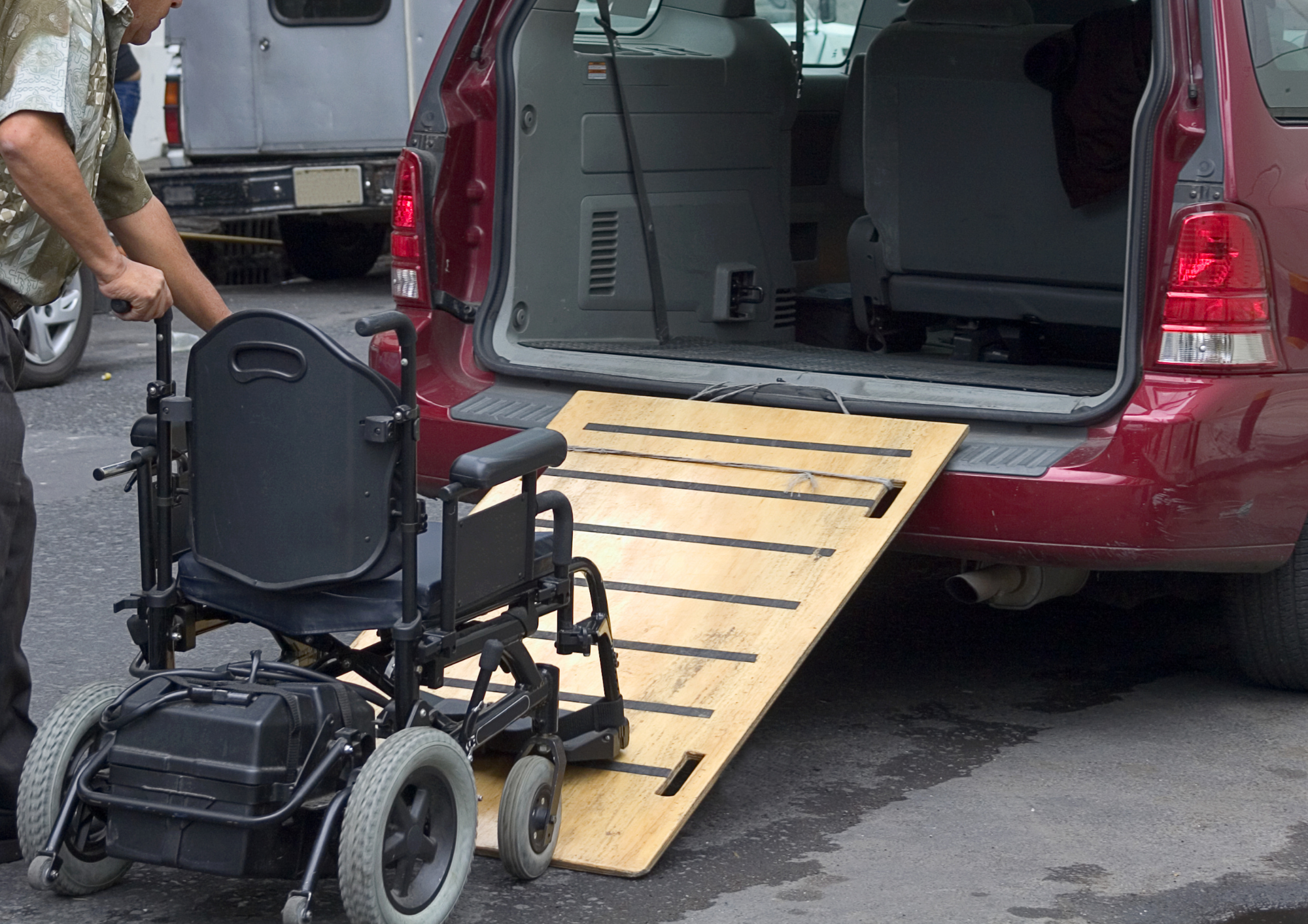A person in a mobility scooter is loading it onto the back of a red vehicle using a wooden ramp. The vehicle's trunk is open, and the ramp is extended from the trunk to the ground.