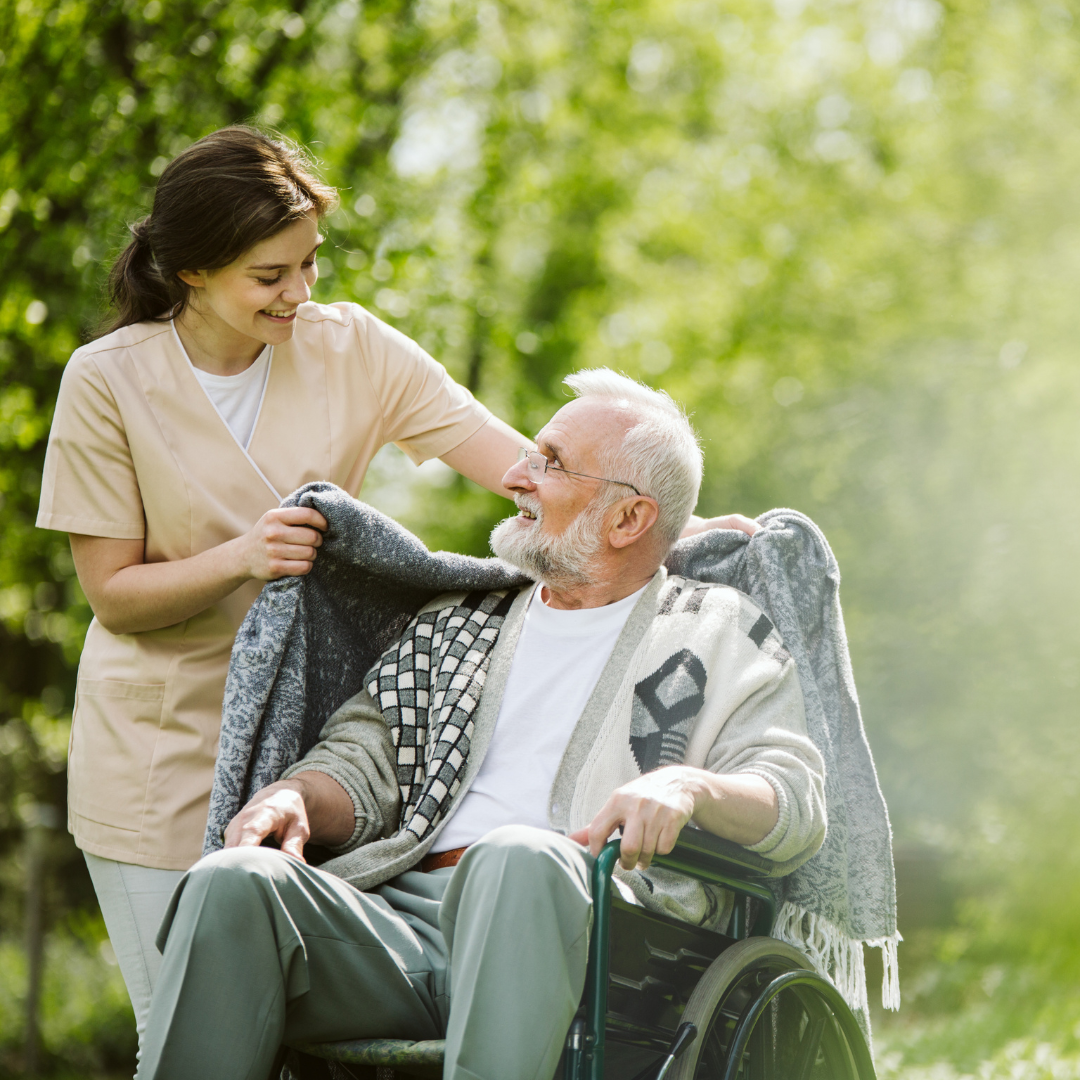 A young female nurse or caregiver smiling and talking to an elderly man in a wheelchair outdoors in a green park, with sunlight filtering through trees.