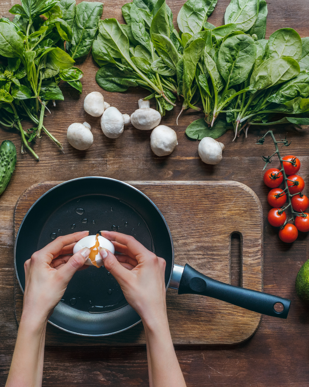 Person cracking an egg into a frying pan on a wooden cutting board, surrounded by fresh basil, spinach, cherry tomatoes, mushrooms, cucumber, and avocado.