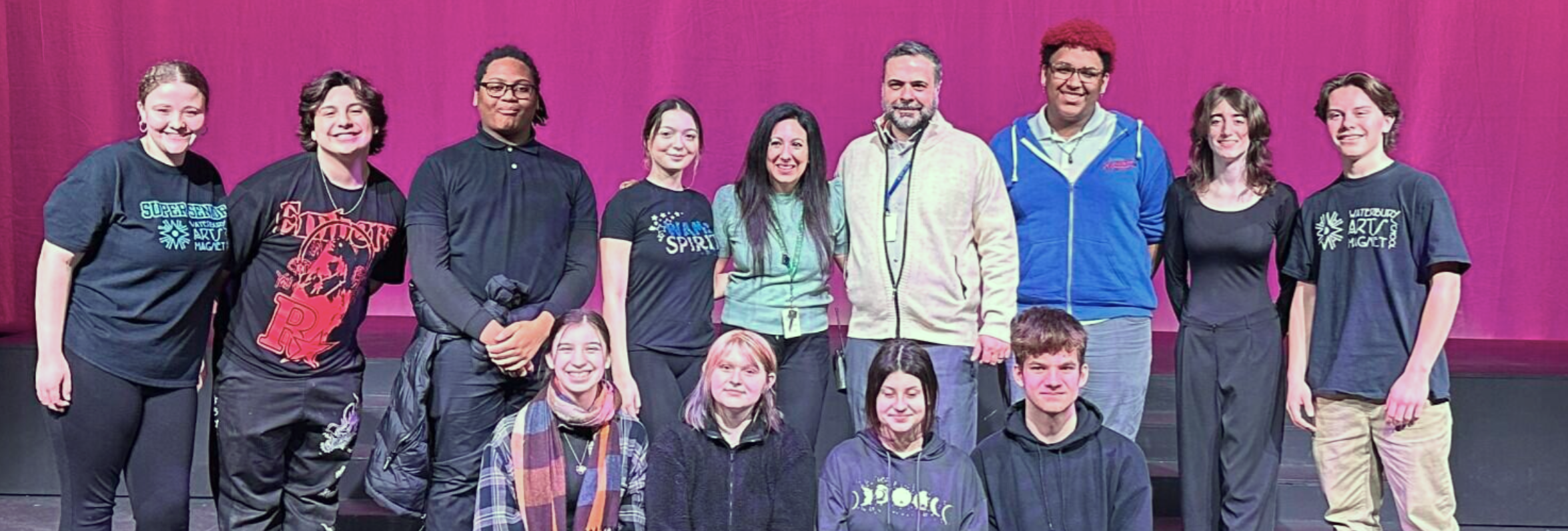 Group of people standing on stage in front of a pink curtain, some wearing t-shirts and others wearing jackets or hoodies, smiling at the camera.