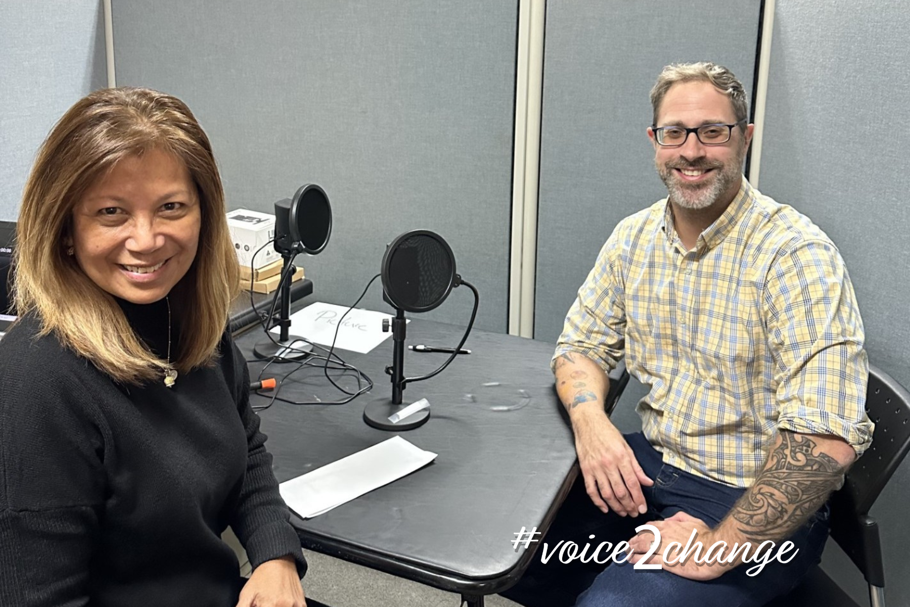 A woman and a man sit at a table with microphones in a recording studio or podcast room, smiling at the camera. The woman has shoulder-length light brown hair, wearing a black top, and a necklace. The man has short light brown hair, glasses, a beard, and tattoos on his arms, wearing a yellow plaid shirt. There are papers, a computer monitor, and audio equipment on the table. The text #voice2change is in the bottom right corner.