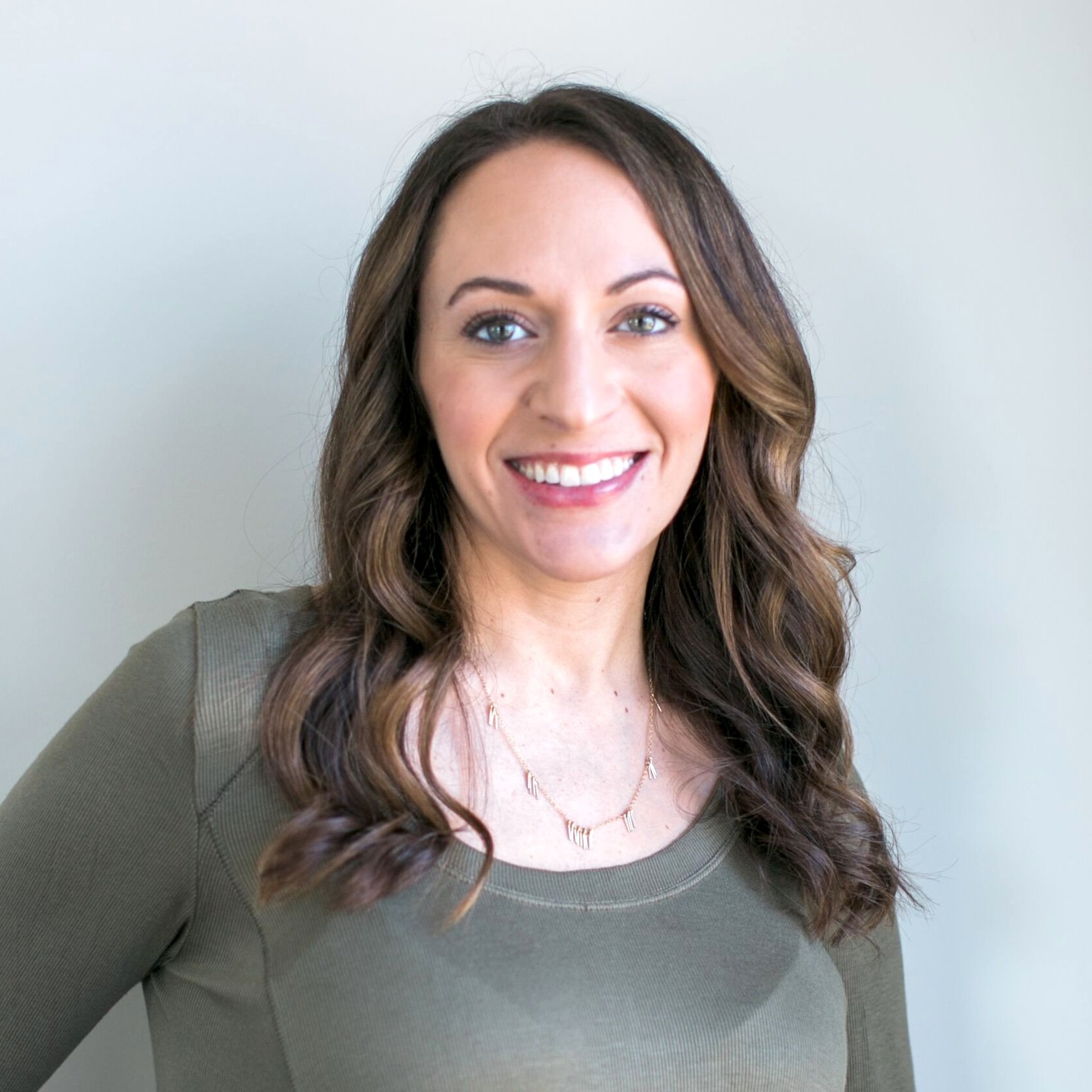 A woman with long, wavy brown hair and a bright smile, wearing a green top and a delicate gold necklace, standing against a light-colored background.