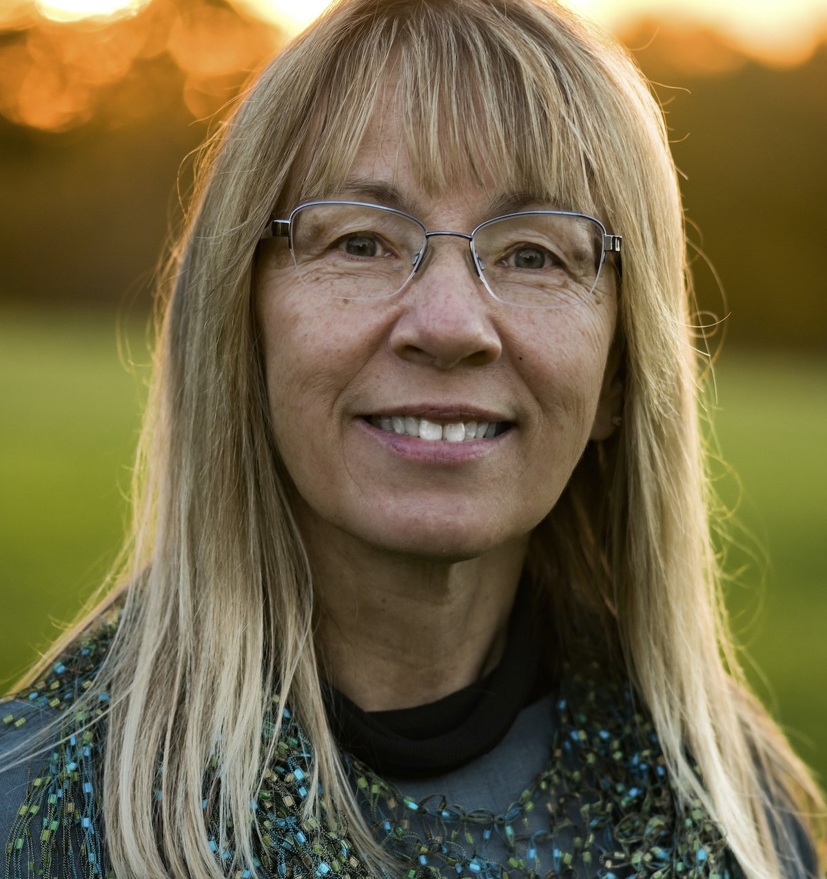 Close-up of a smiling woman with long blonde hair, wearing glasses and a black scarf, outdoors during sunset with a blurred green field in the background.