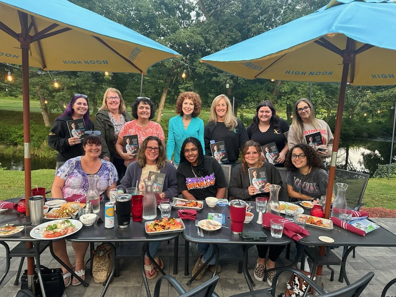 A group of women gathered outdoors at a dining table under a large yellow and blue umbrella, with a scenic background of trees and a pond. They are smiling and holding copies of a book, with plates of food and drinks on the table.