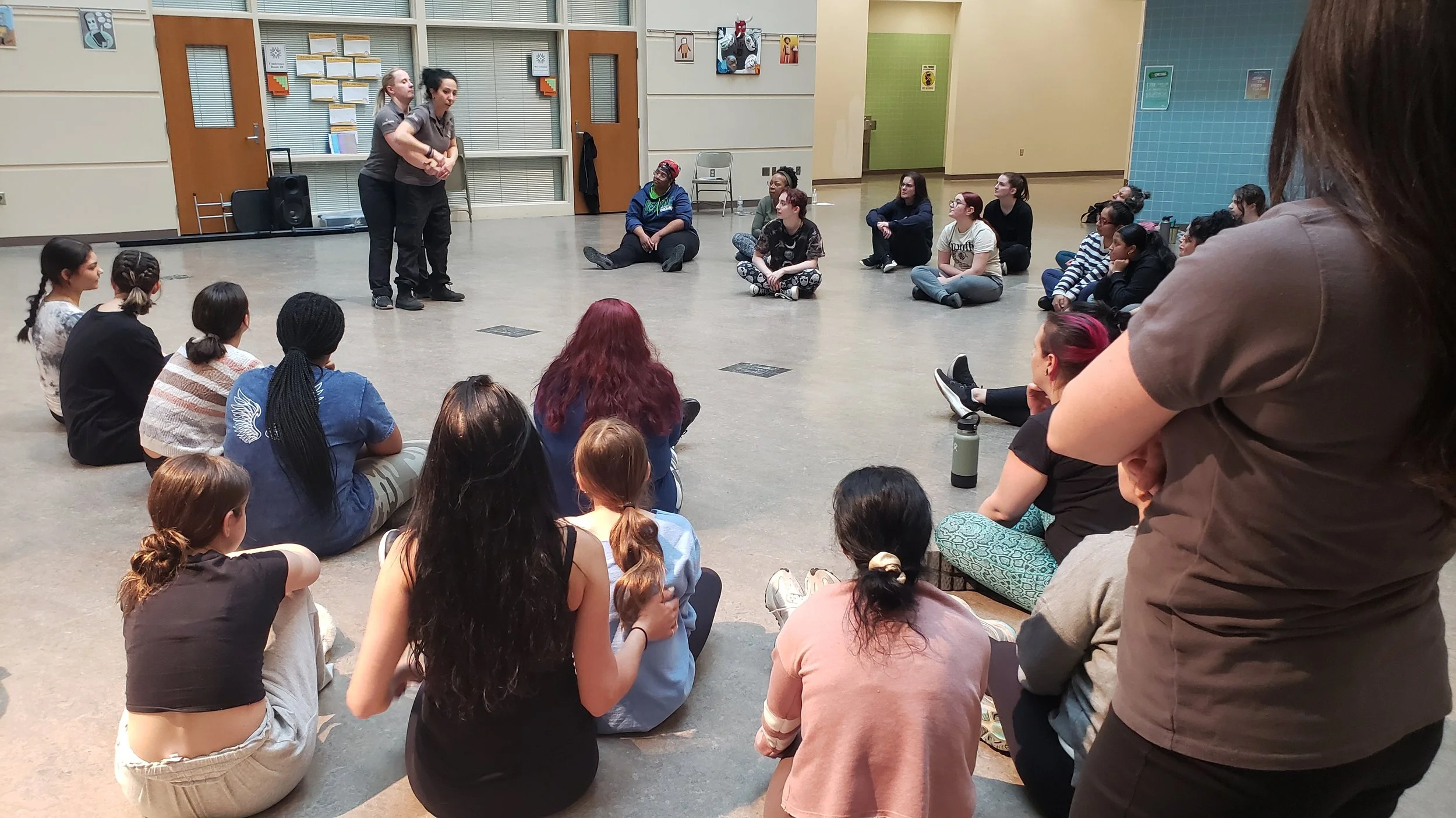 A group of young women sitting on the floor in a semi-circle in a large room, watching a performance or presentation by two young women standing in front of them. The room has colorful walls, posters, and a door with a window.
