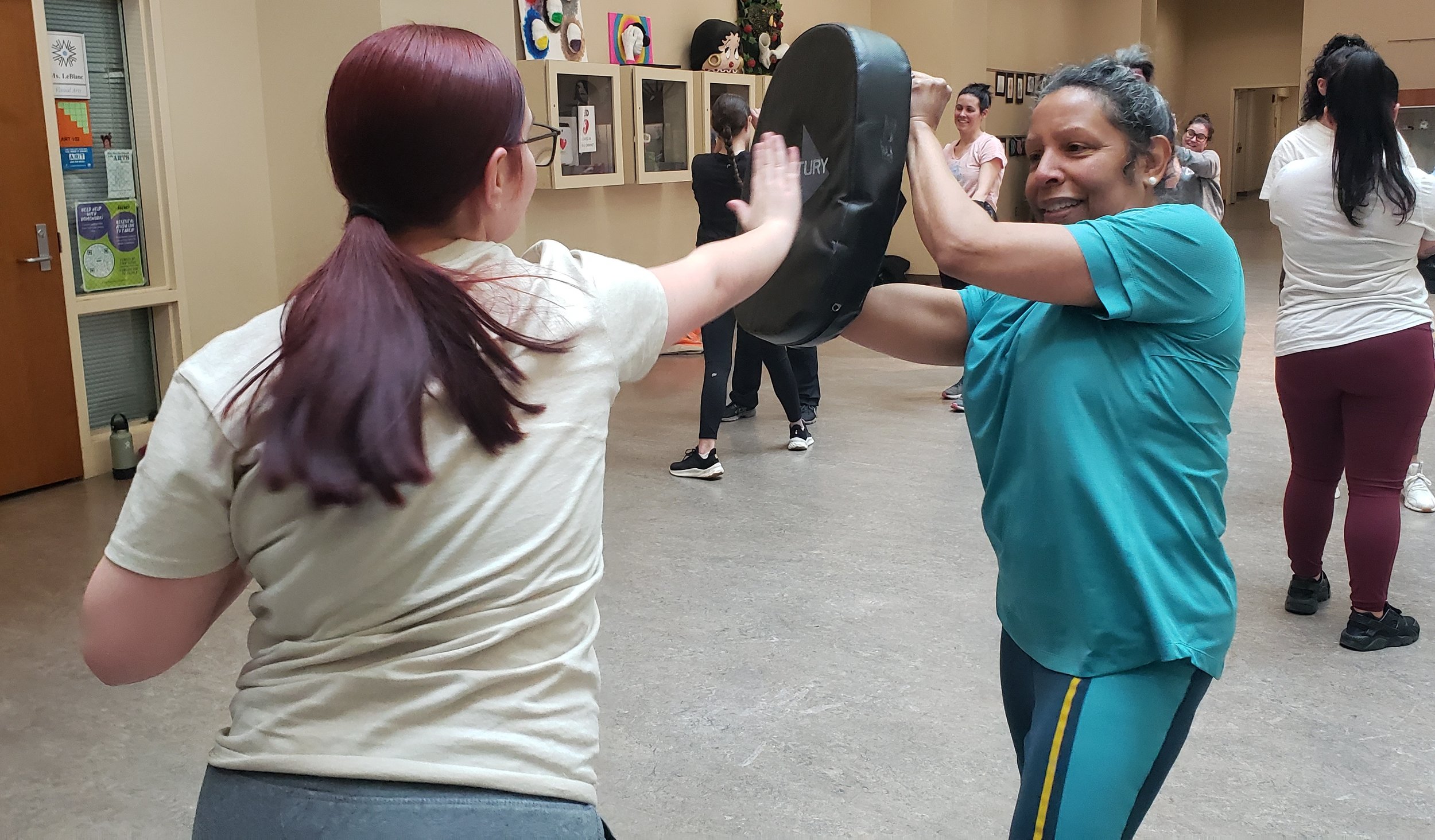 Two women practicing martial arts in a gym, with other women in the background. One woman is holding a padded shield while the other is performing a strike.
