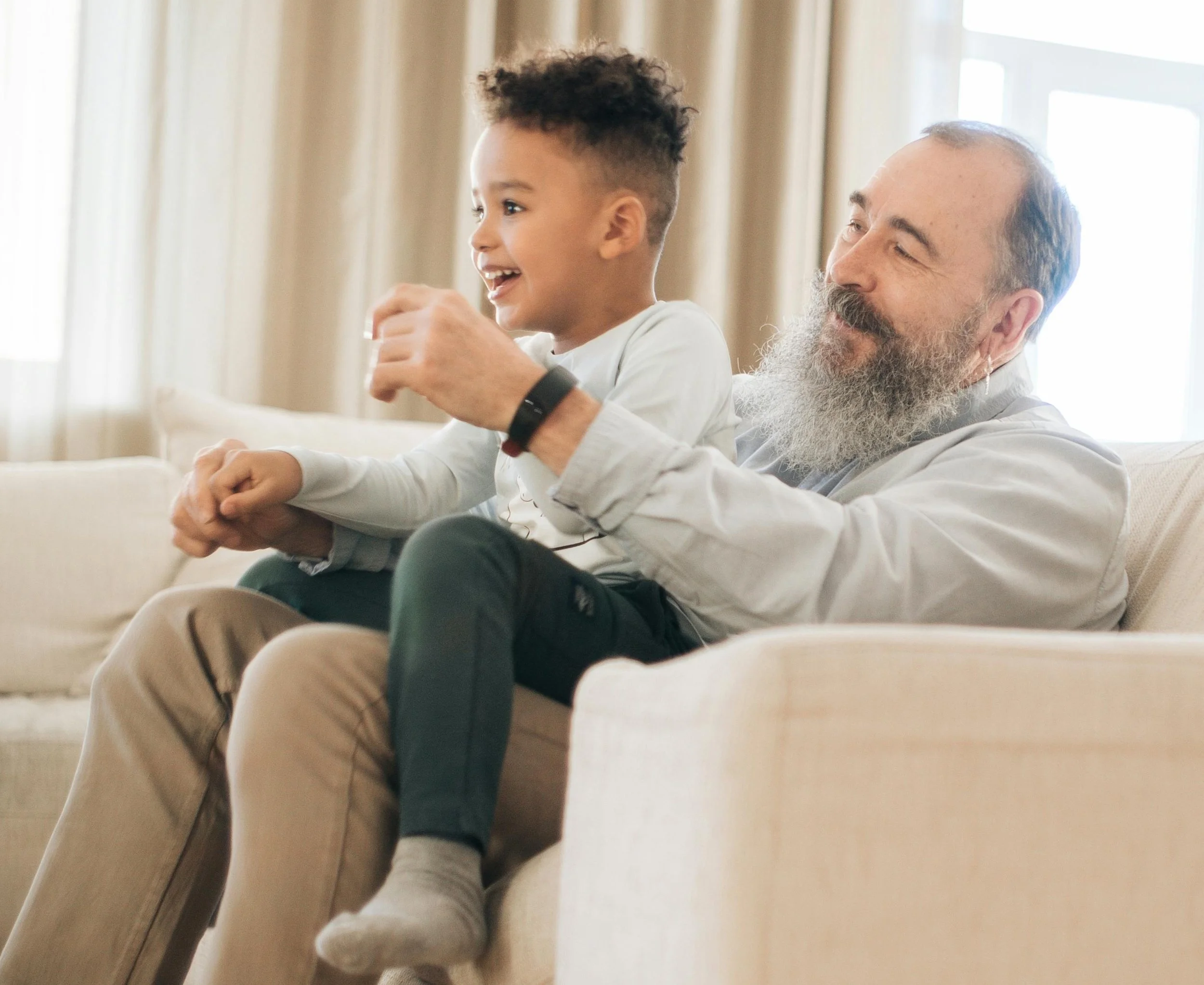 A young boy and an older man, possibly his grandfather, sitting on a beige couch in a well-lit living room, enjoying a happy moment together.