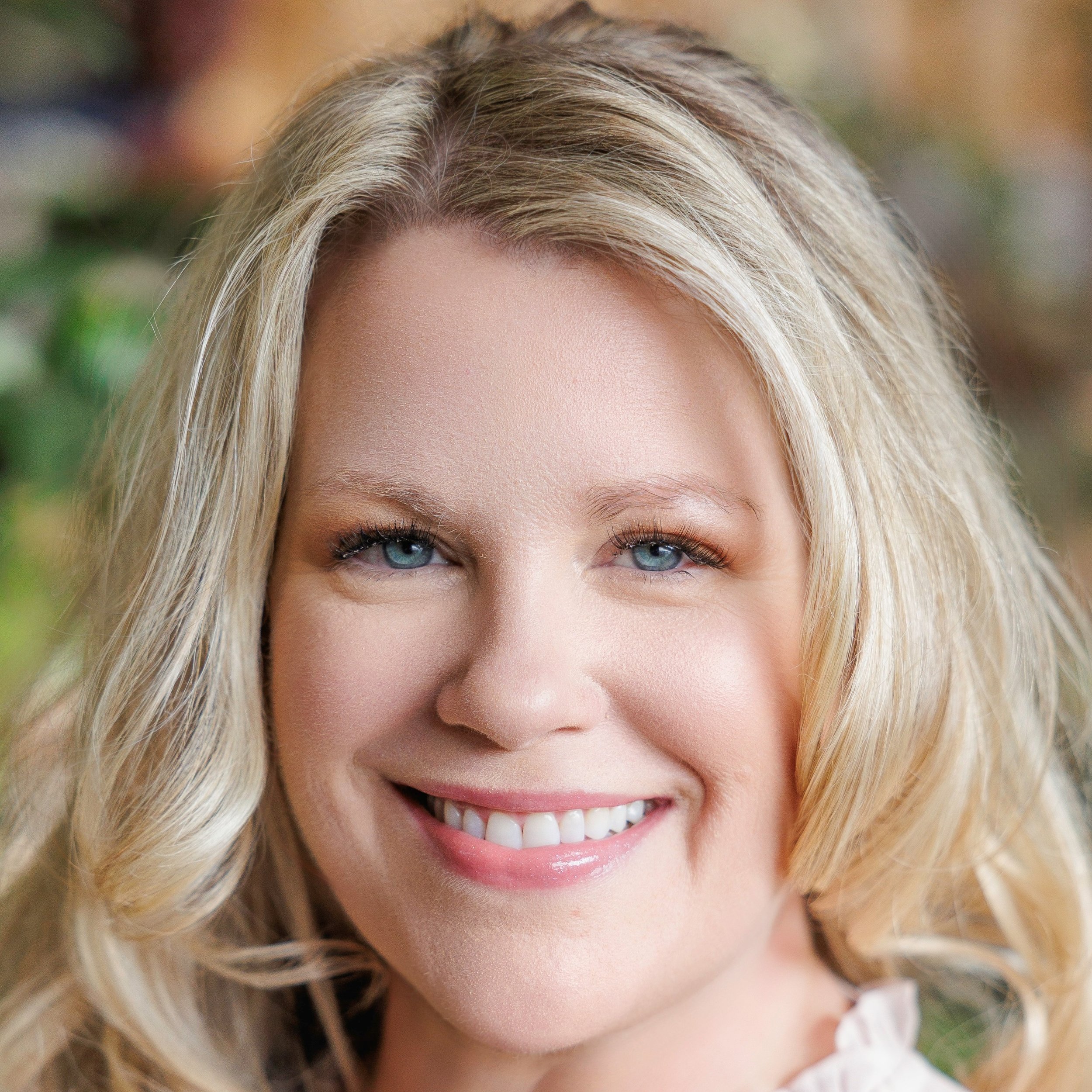 Close-up of a smiling blonde woman with blue eyes, outdoors with blurred greenery in the background.
