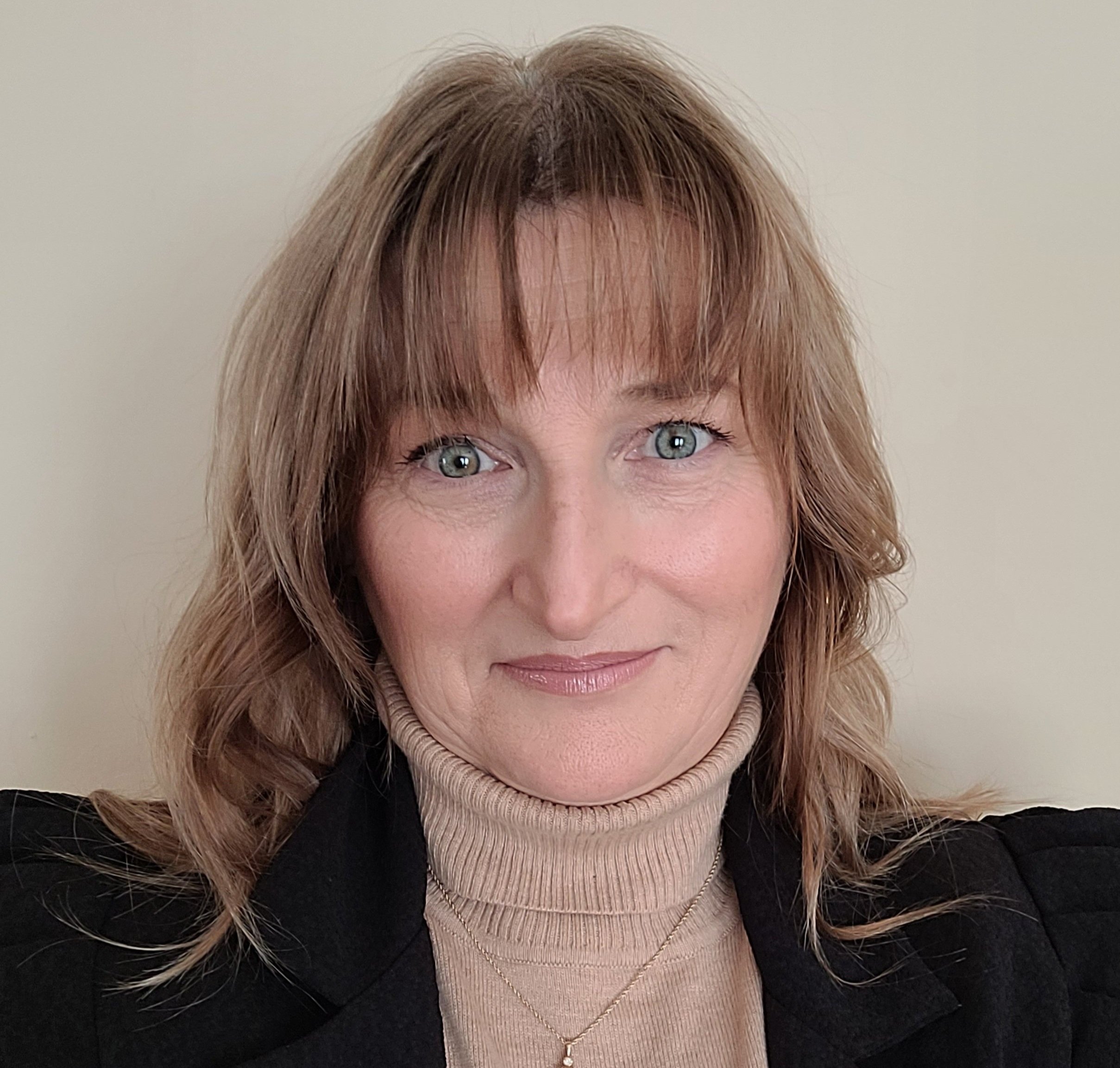 Close-up portrait of a woman with shoulder-length wavy light brown hair, wearing a black blazer and a beige turtleneck. She is smiling slightly and looking directly at the camera against a plain light-colored background.