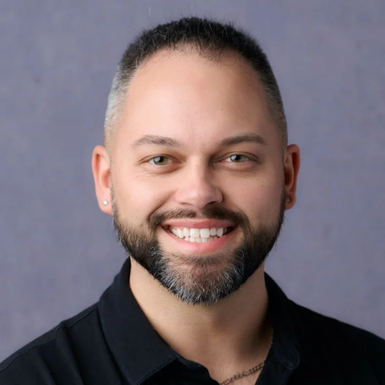 Close-up portrait of a smiling man with a beard and short hair, wearing a black shirt, against a gray background.