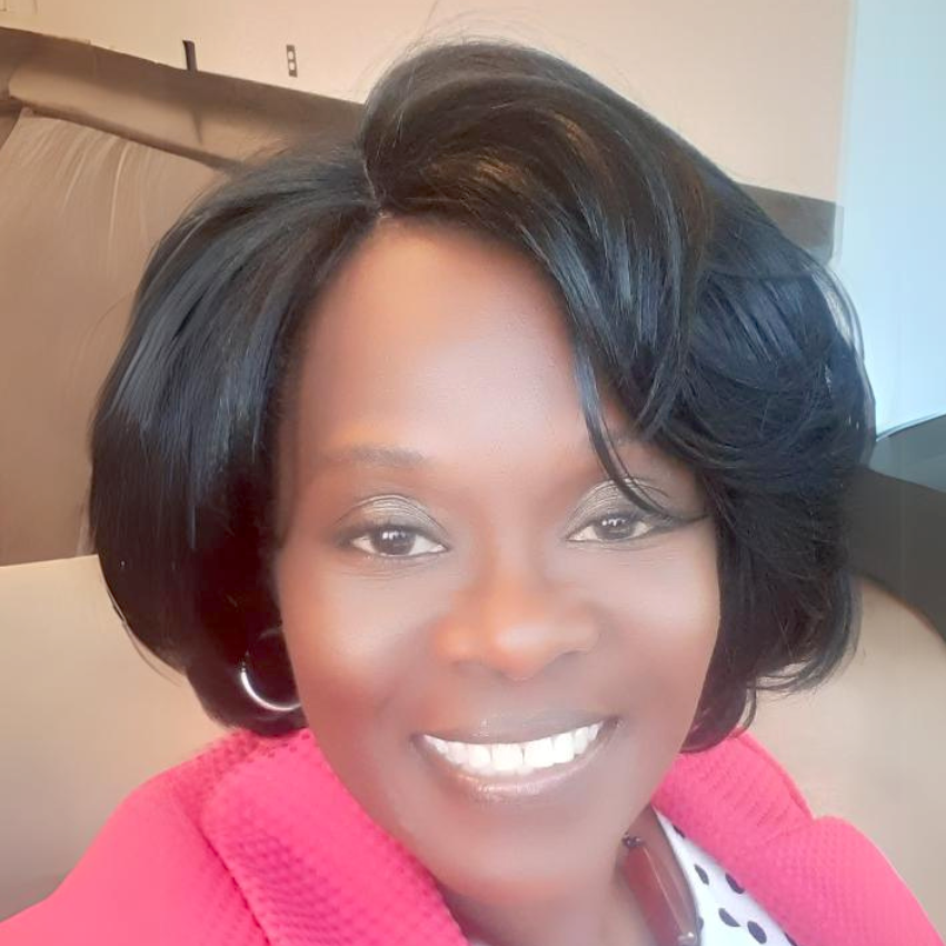 A woman with short black hair styled in waves, wearing a pink blazer and a white and black polka dot top, smiling indoors.