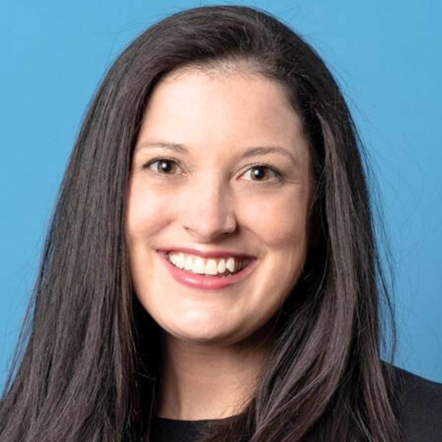 A woman with long brown hair smiling, wearing a black top, against a blue background.