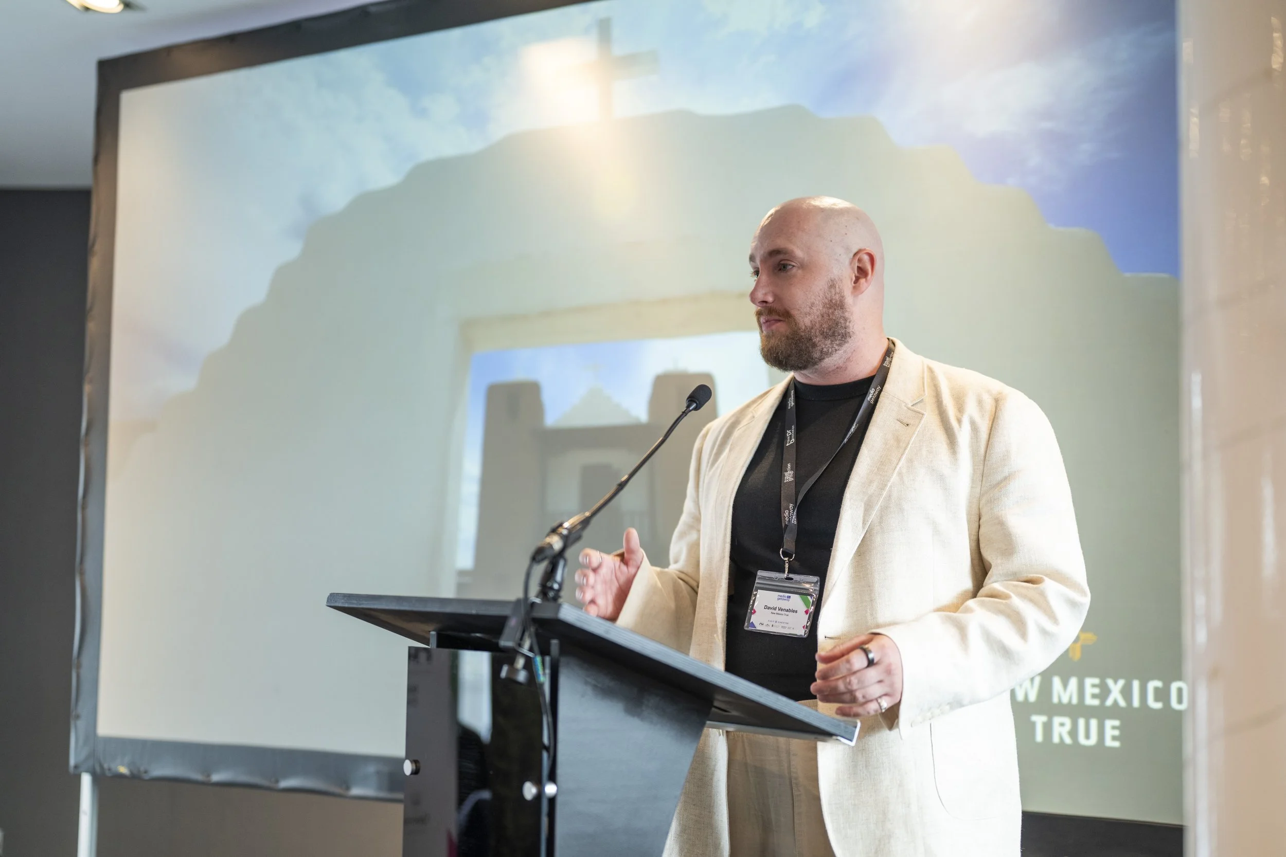 Man with a bald head and beard giving a presentation at a podium in front of a projection screen displaying a scenic background and text that reads 'New Mexico True.'