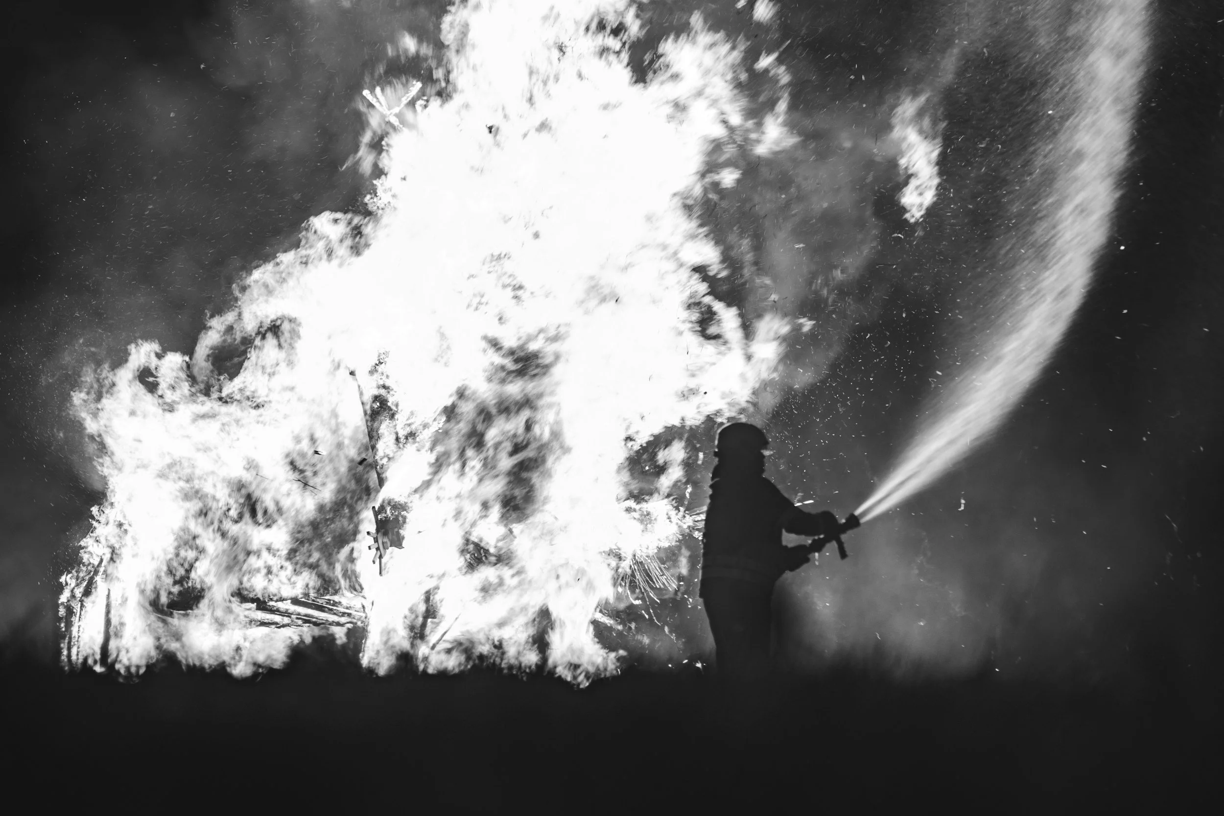 A person in a helmet holding a fire hose, spraying water onto a large fire with flames and smoke in the background, in black and white.
