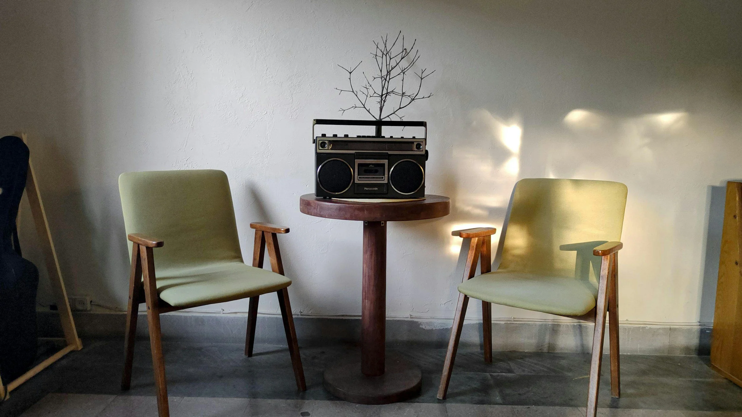 Interior of a room with two green chairs with wooden armrests facing a small round wooden table. On the table, there's a vintage boombox with a small leafless decorative tree behind it. Light creates a bright pattern on the wall in the background.