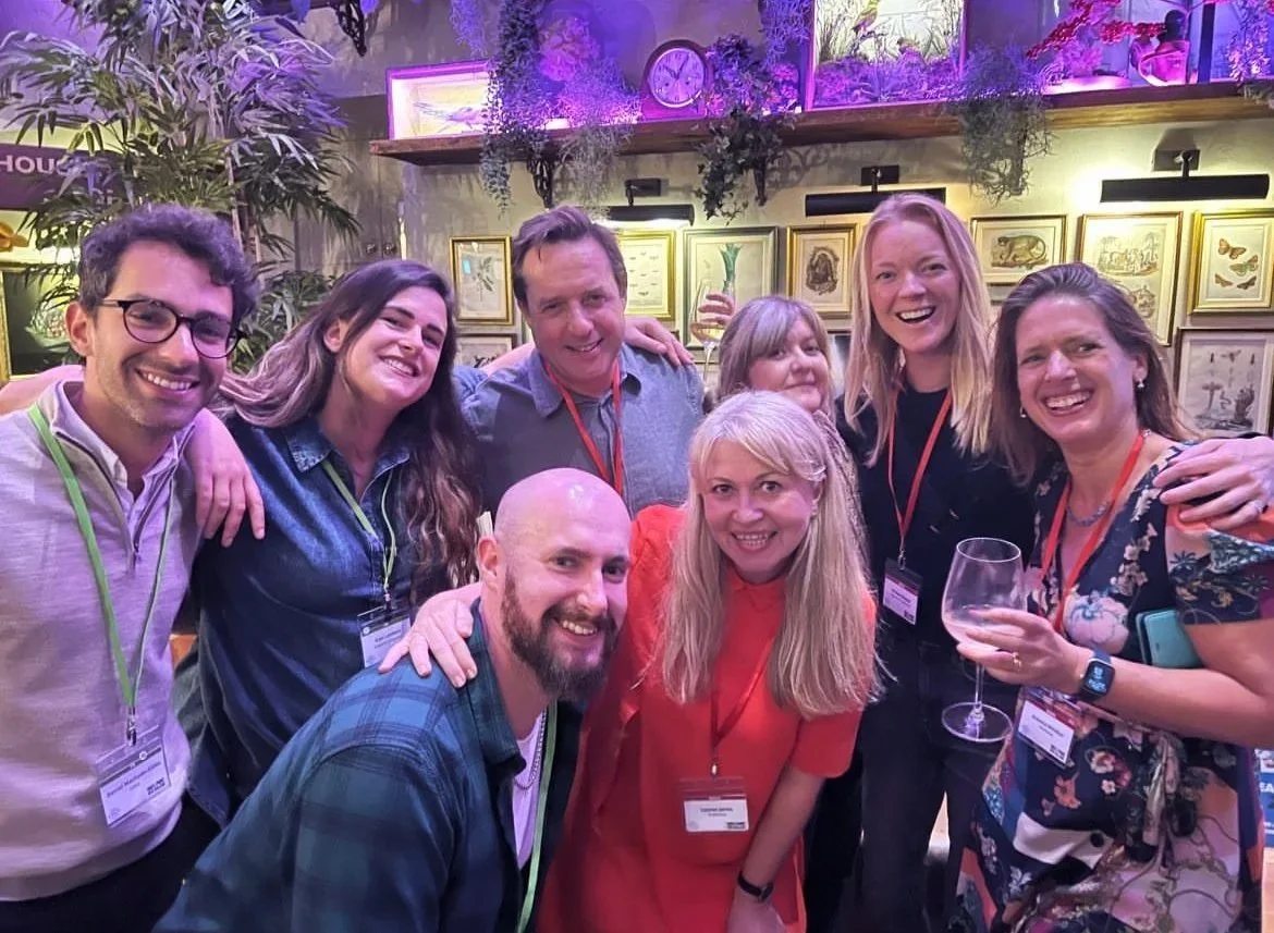 Group of eight smiling people posing for a photo at an indoor event or gathering, some holding glasses of wine, with a colorful, decorated background including framed artwork, plants, and purple lighting.