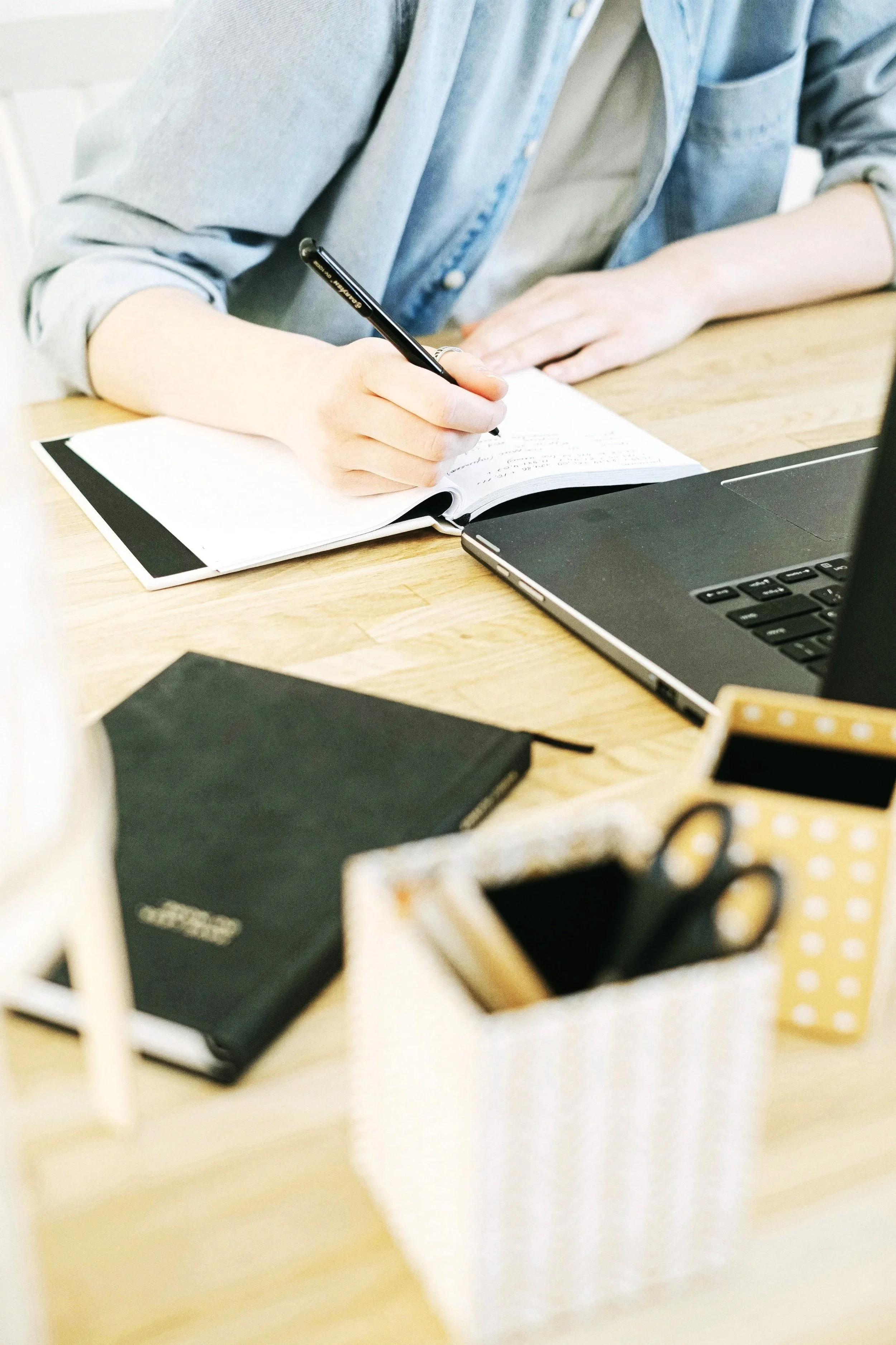 Person writing in a notebook at a desk with a laptop, closed notebook, and various office supplies.