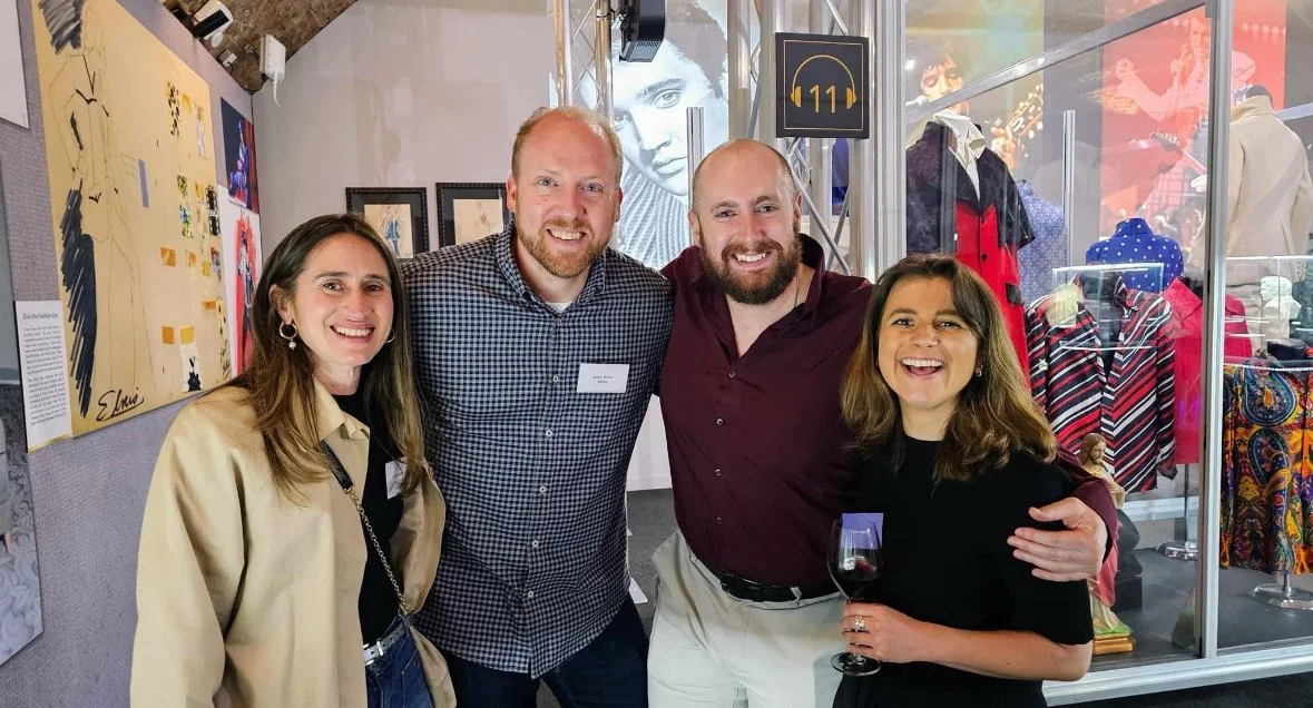 Four people smiling and posing together at an art exhibit or gallery, with artwork on the wall behind them and a display case with colorful clothing in the background. One woman is holding a glass of red wine.