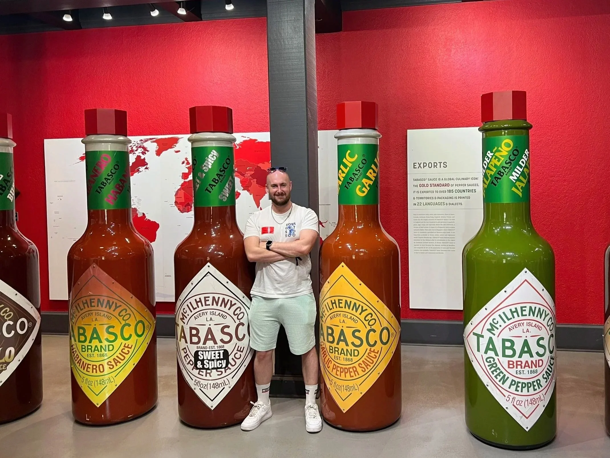 A man standing next to large bottles of Tabasco hot sauce in a display at a museum or exhibit. The bottles are labeled with different flavors and are much larger than a typical bottle.