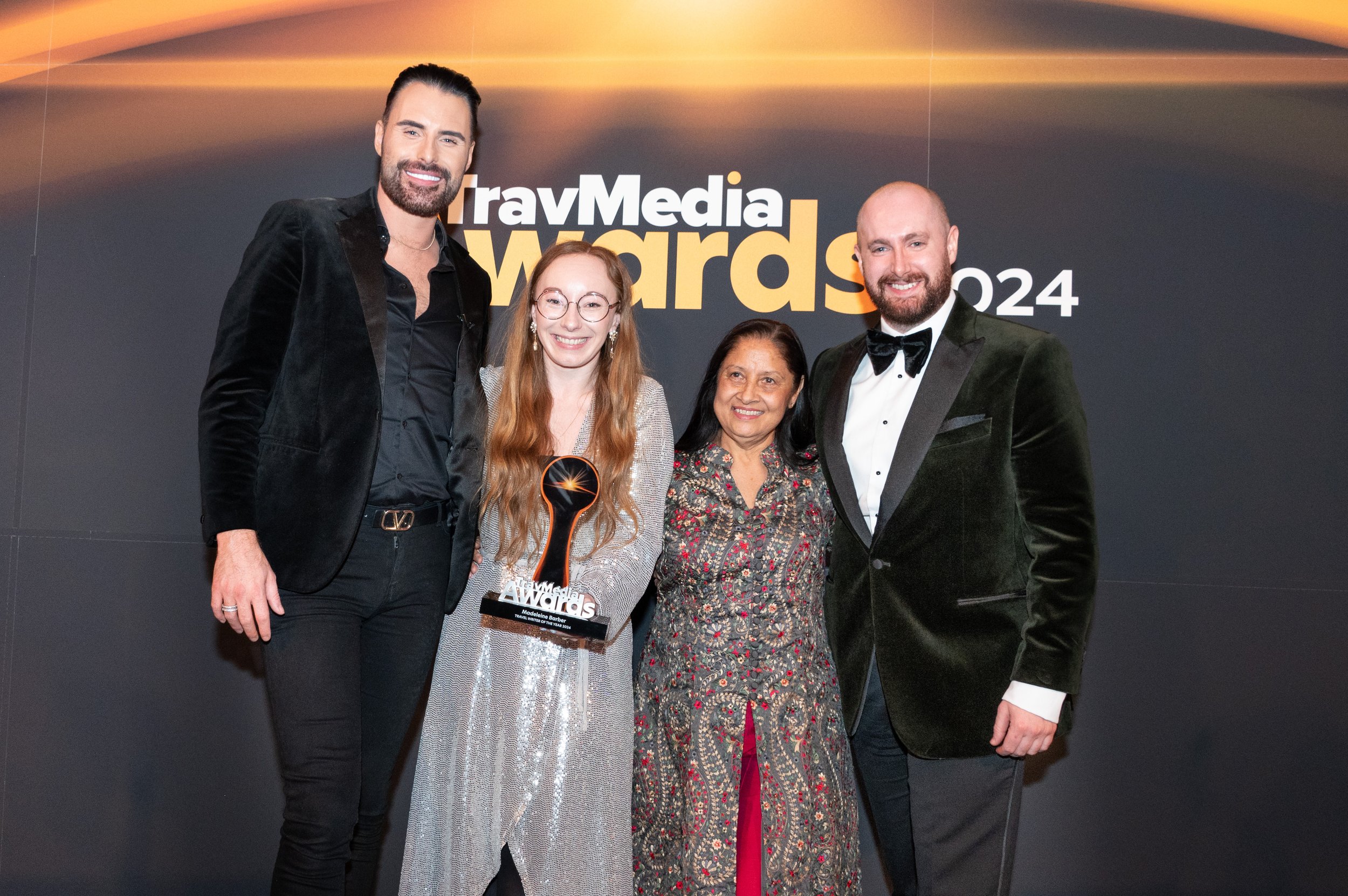 A group of five people dressed in formal attire posing at the TravMedia Awards 2024 event, with a woman holding an award trophy.