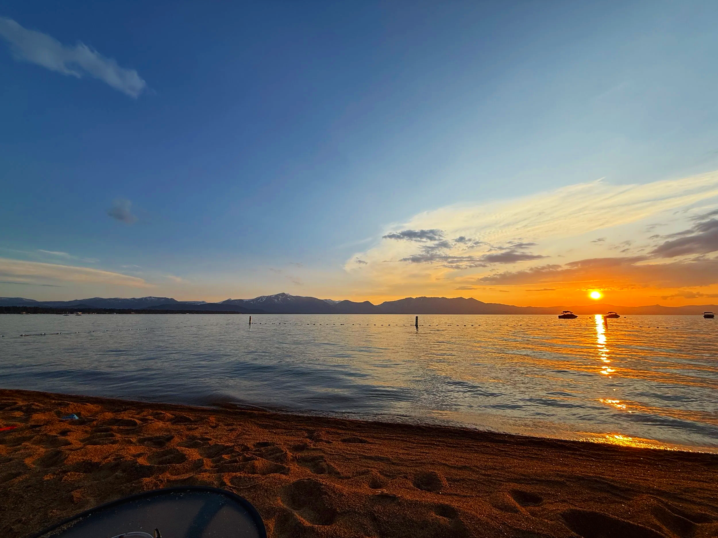 Sun setting over a calm lake with boats, mountains in the background, and a sandy beach in the foreground.