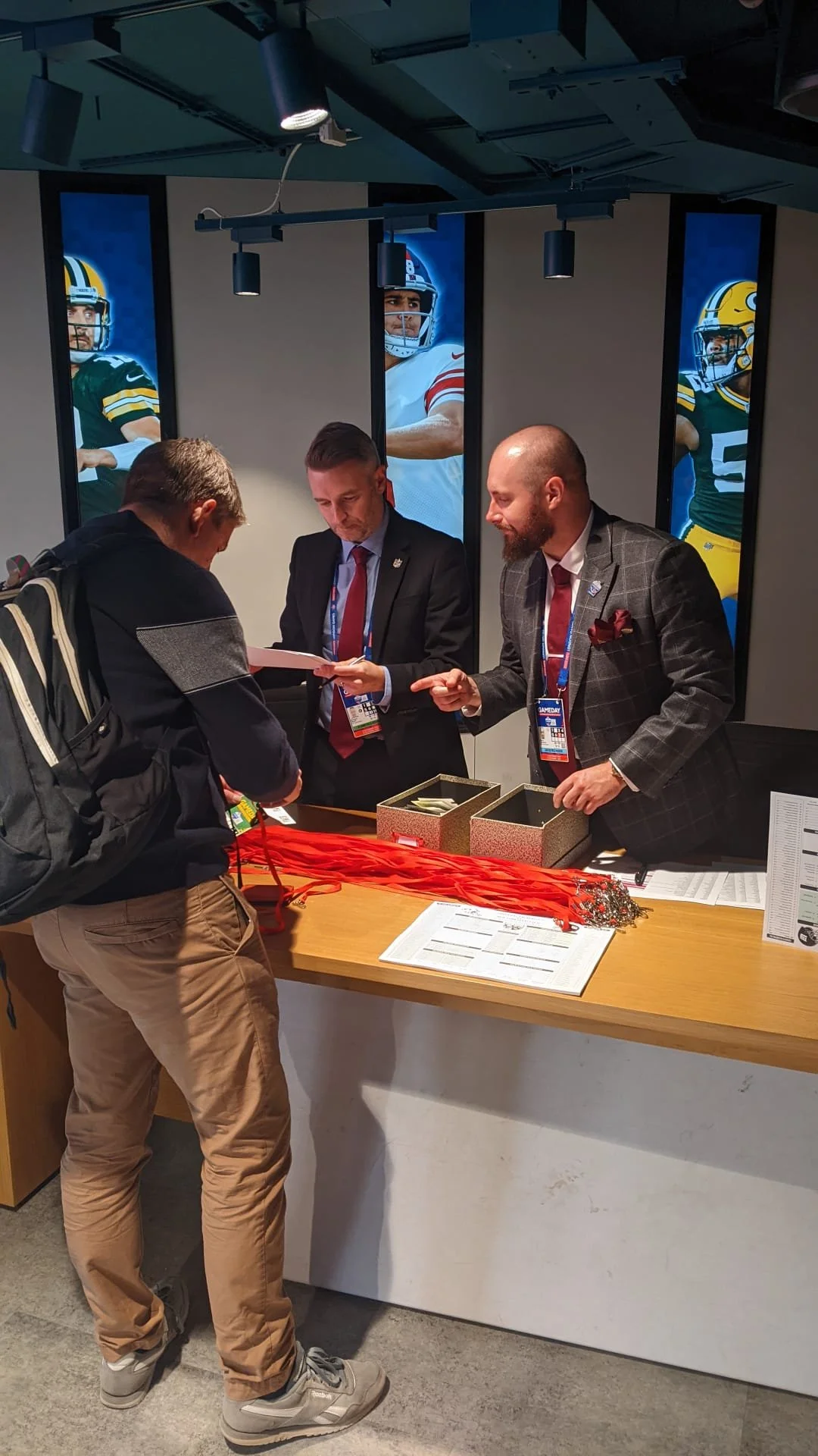 A young man with a backpack at a registration desk speaking with two men in suits, all wearing conference badges, in an indoor setting. Behind them are large images of football players on a wall.