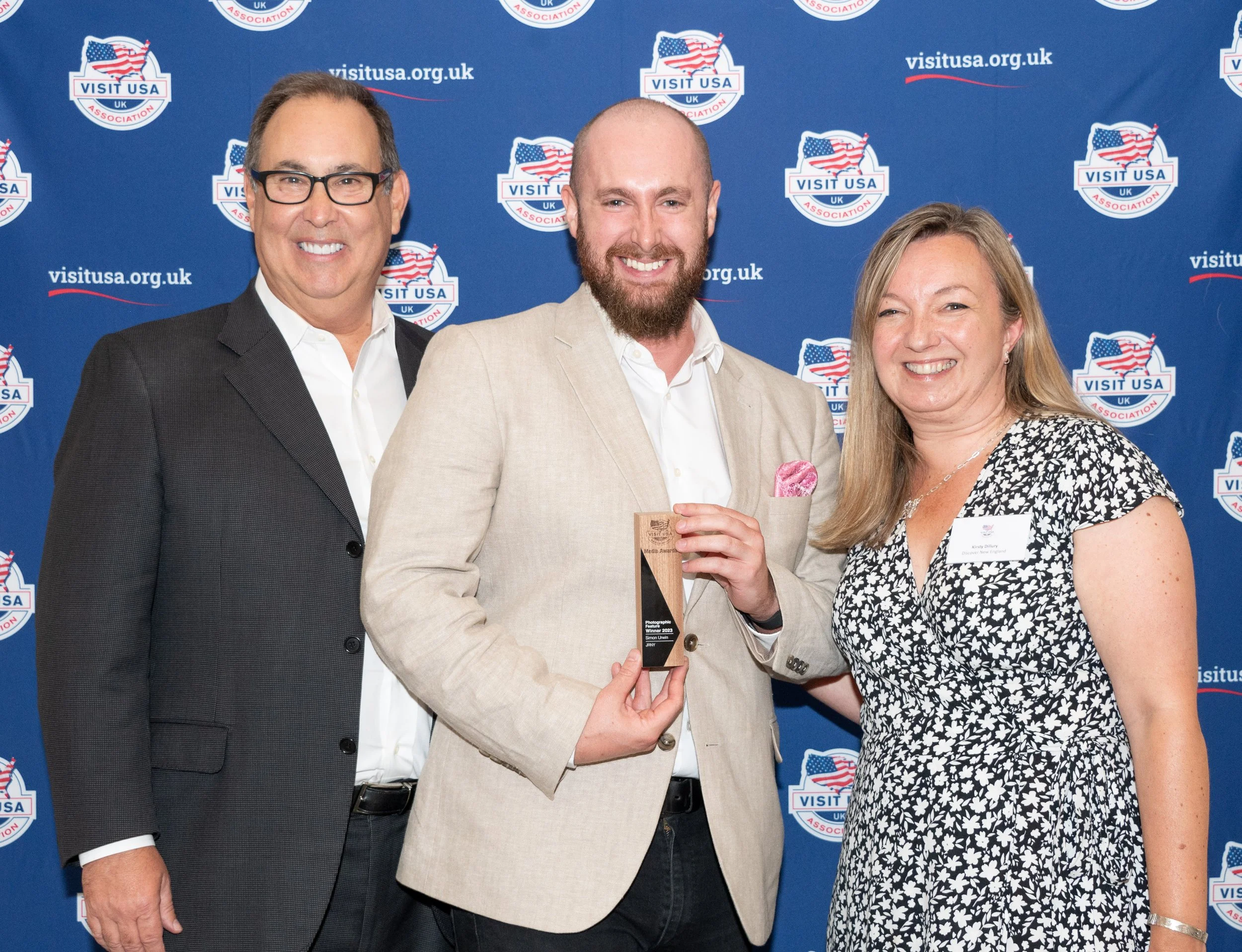 Three people standing in front of an event backdrop with a 'Visit USA UK' logo, smiling. The man in the middle is holding a trophy and wears a beige blazer and pink pocket square. The woman on the right wears a black and white floral dress, and the man on the left wears a dark suit and glasses.