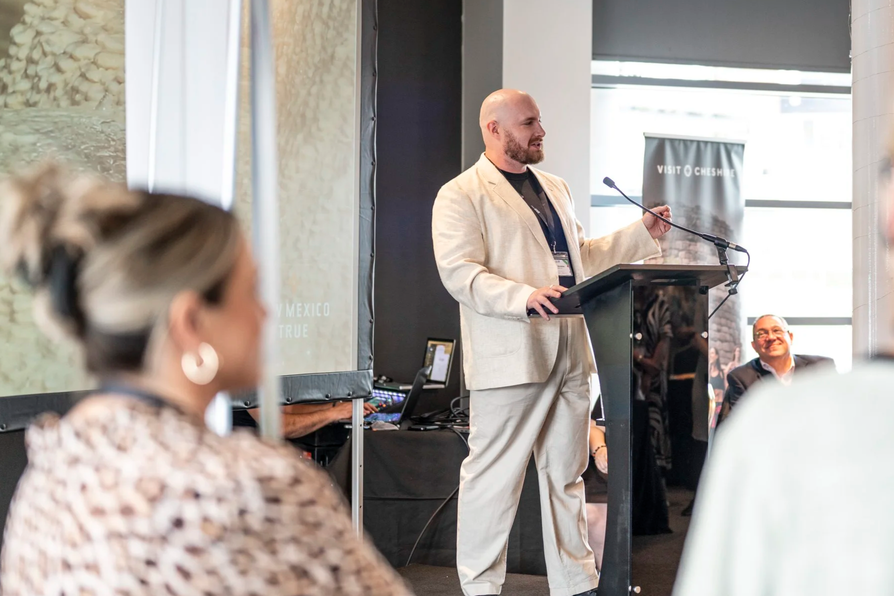 A man with a beard in a beige suit is giving a speech at a conference. He is standing behind a podium with a microphone, speaking to the audience. There is a large screen behind him and people sitting and listening attentively.