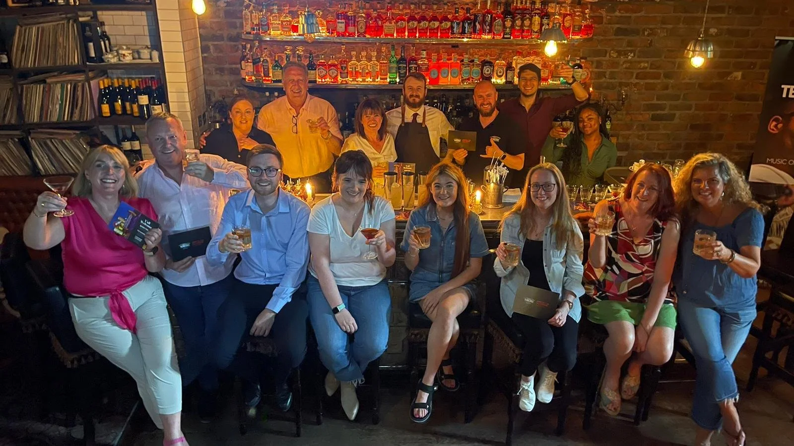 Group of people gathered in a bar or restaurant, smiling and holding drinks, celebrating together with warm lighting and a brick wall background.