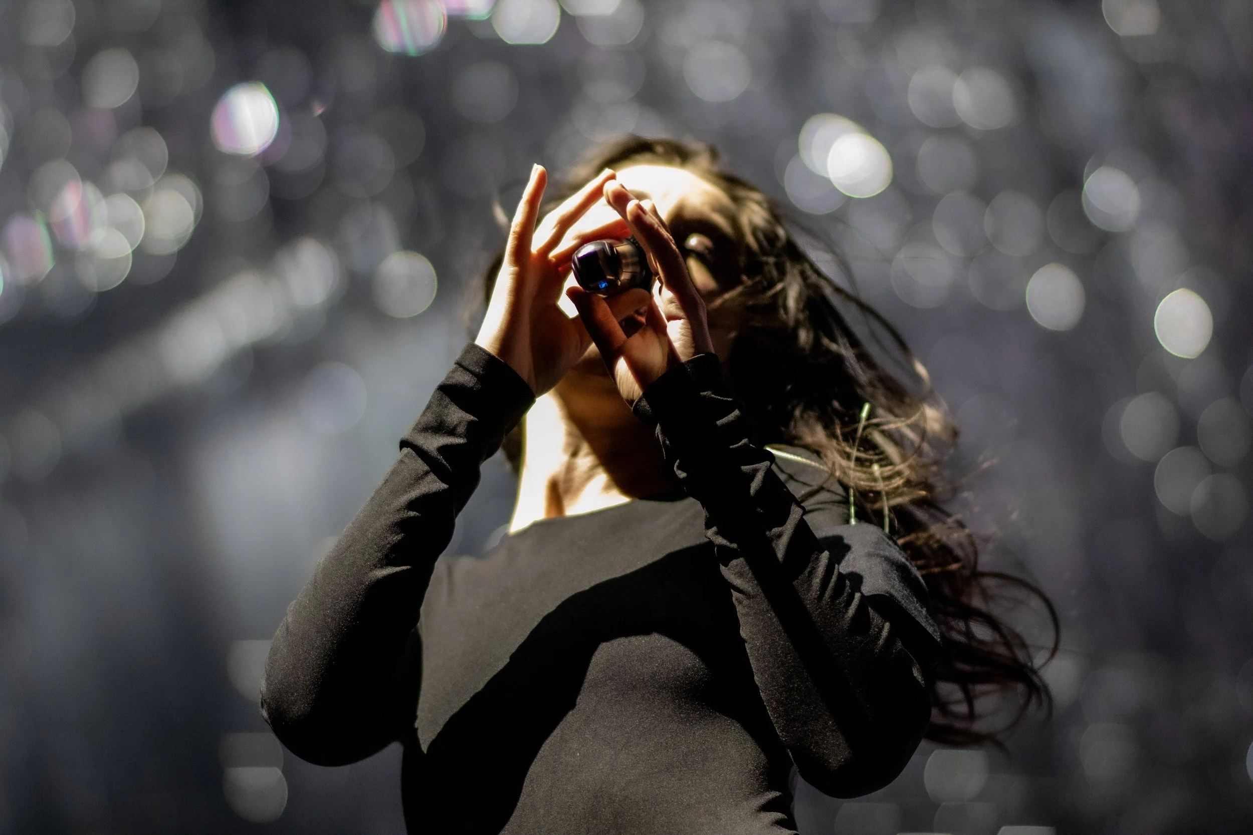 A woman with long dark hair in a black long-sleeve shirt singing or speaking into a handheld microphone on stage with a dark, bokeh background.