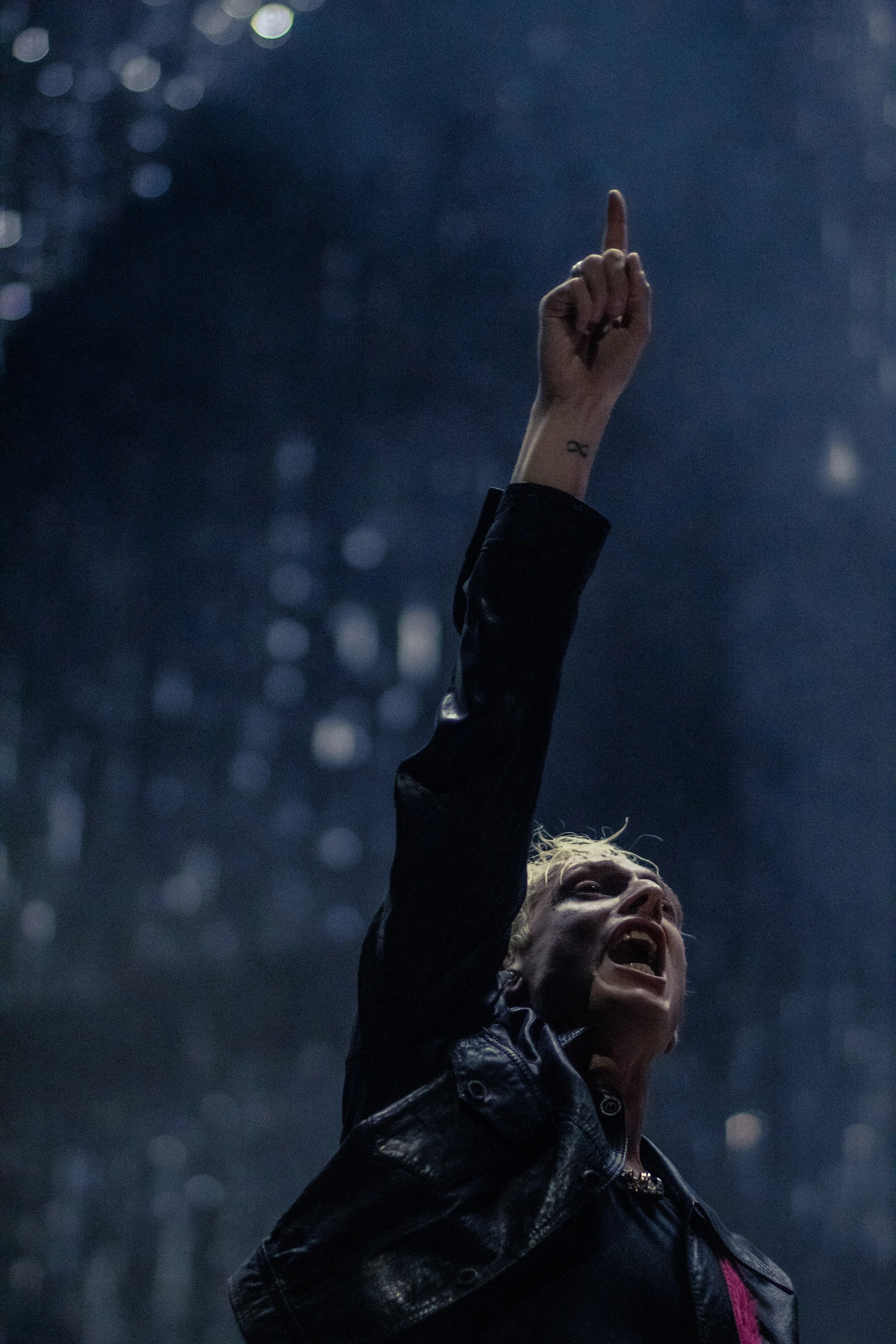 A woman with blonde hair, wearing a black leather jacket, raising her arm with her index finger pointing upward, in a dark, possibly rainy urban setting.