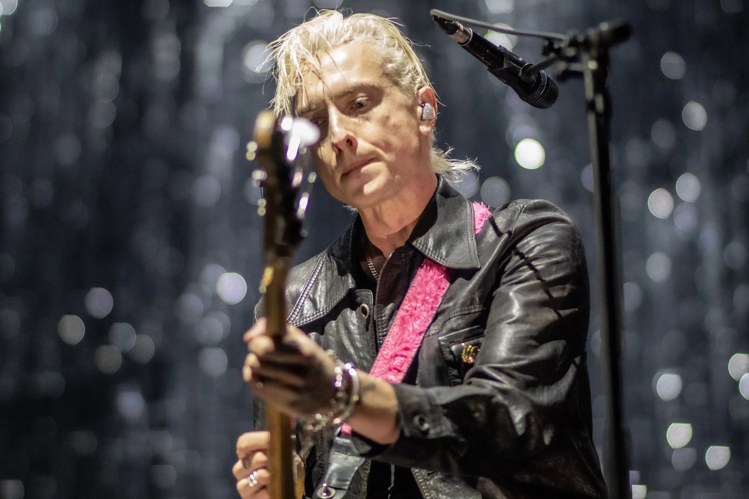 A musician with blond, messy hair wearing a black leather jacket and pink strap playing an electric guitar on stage, with a microphone nearby and a dark, bokeh background.