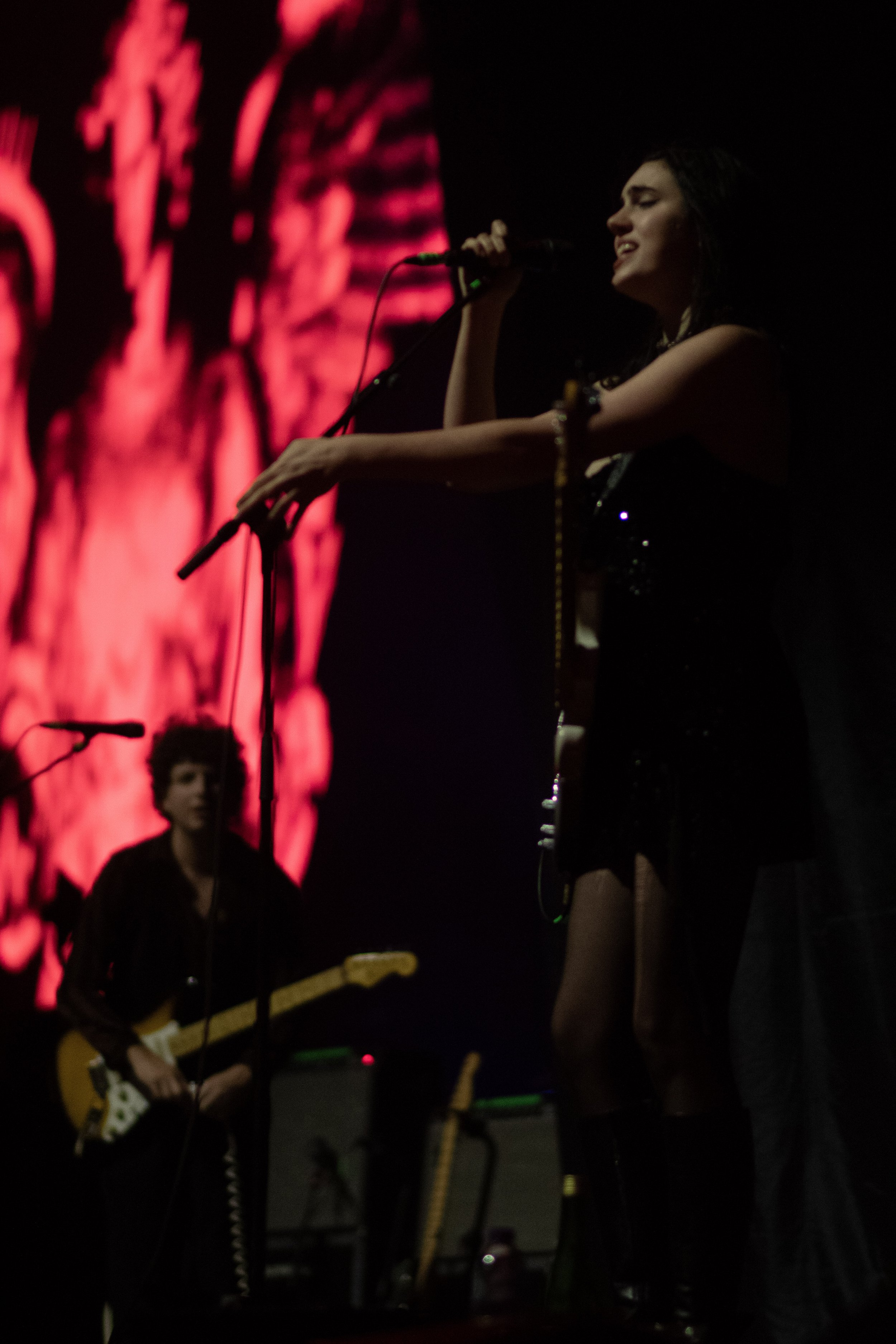 Musicians performing on stage with a red abstract background. A female singer holds a microphone, and a male guitarist plays an electric guitar.