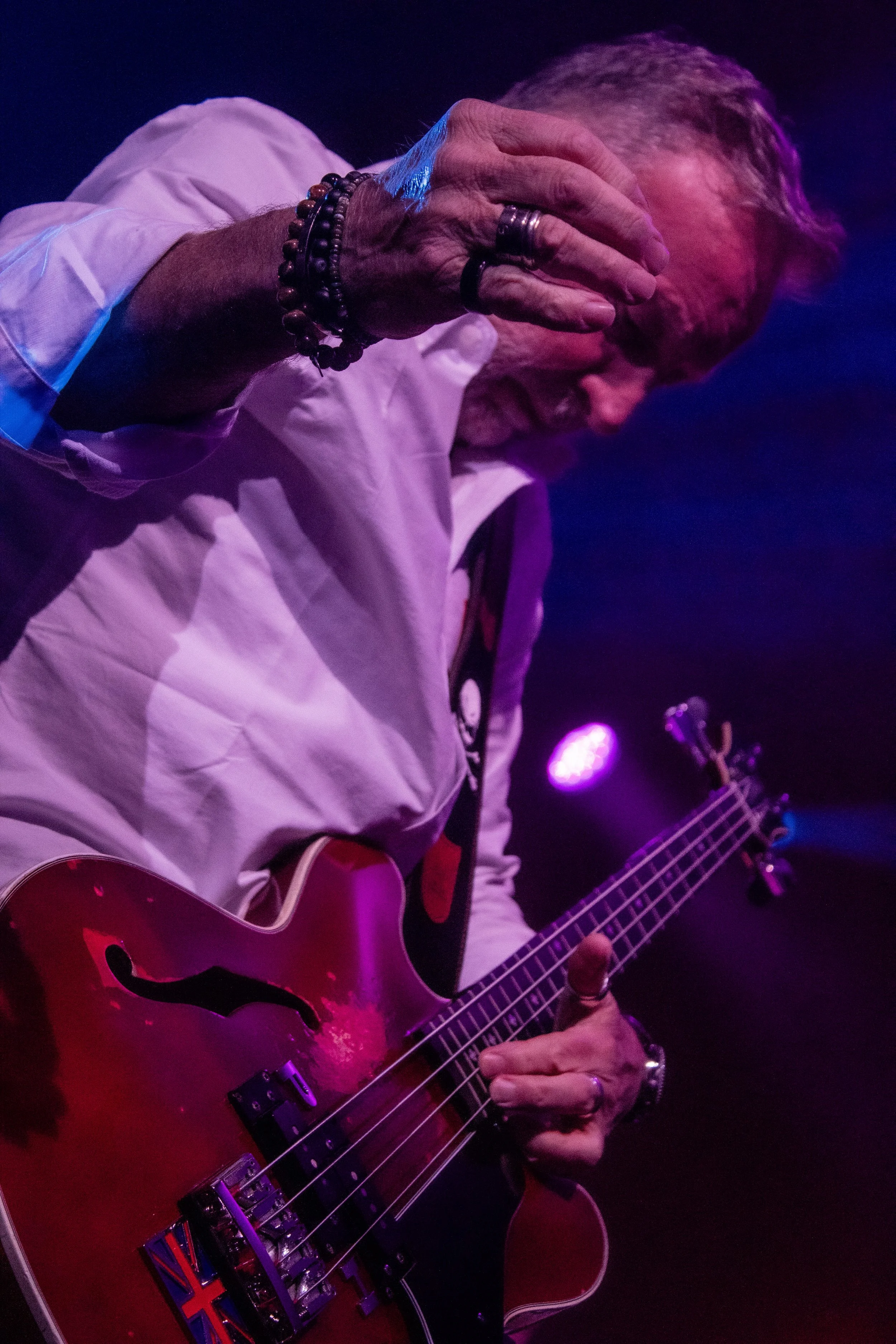A man with gray hair and a beard playing a red electric guitar on stage, wearing a white shirt, surrounded by purple stage lights.