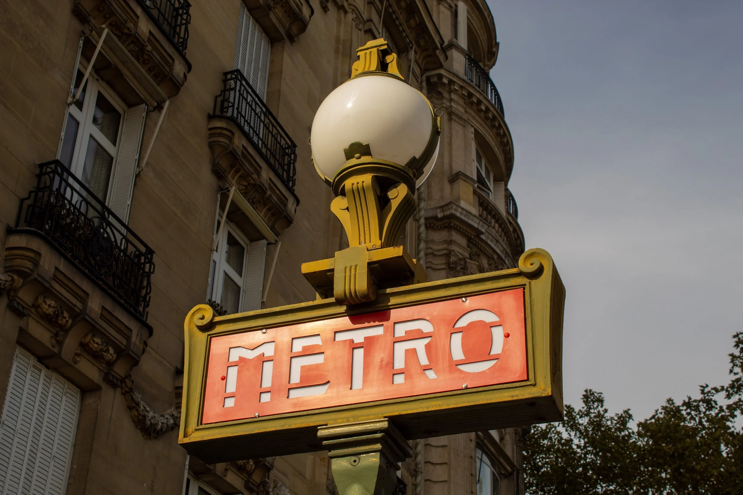 A vintage metro sign with a white globe on top, yellow decorative elements, and a rectangular red background with the word 'METRO' in white. The sign is mounted on a pole in front of a historic European-style building with ornate architectural detail
