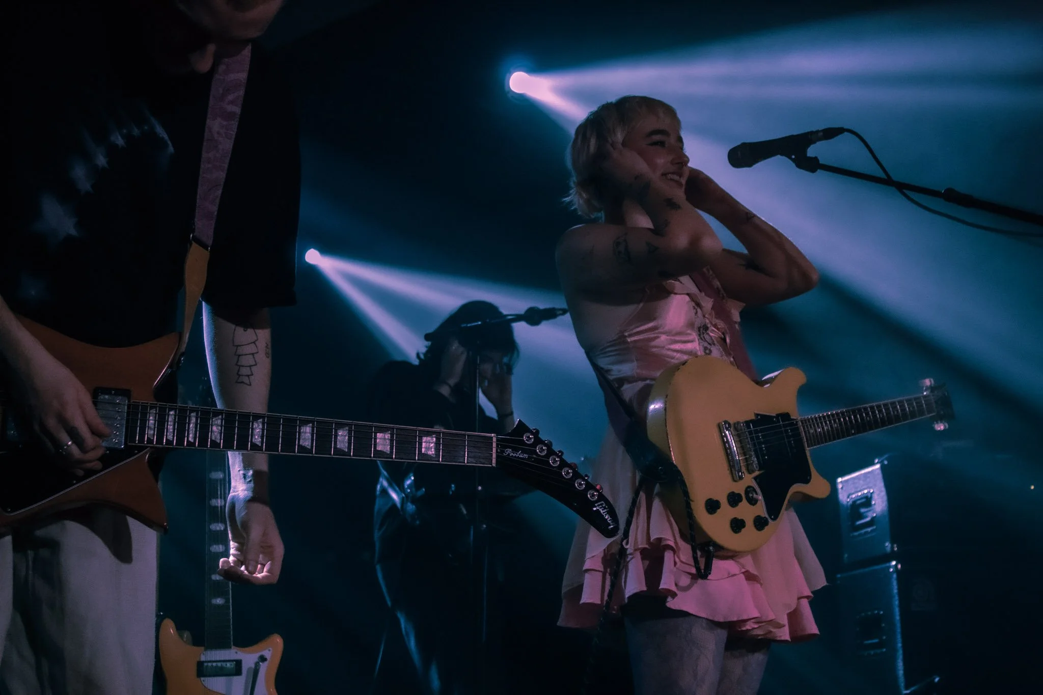 A woman with short blonde hair holding a guitar near a microphone on stage during a performance, with blue stage lighting and a male guitarist next to her.