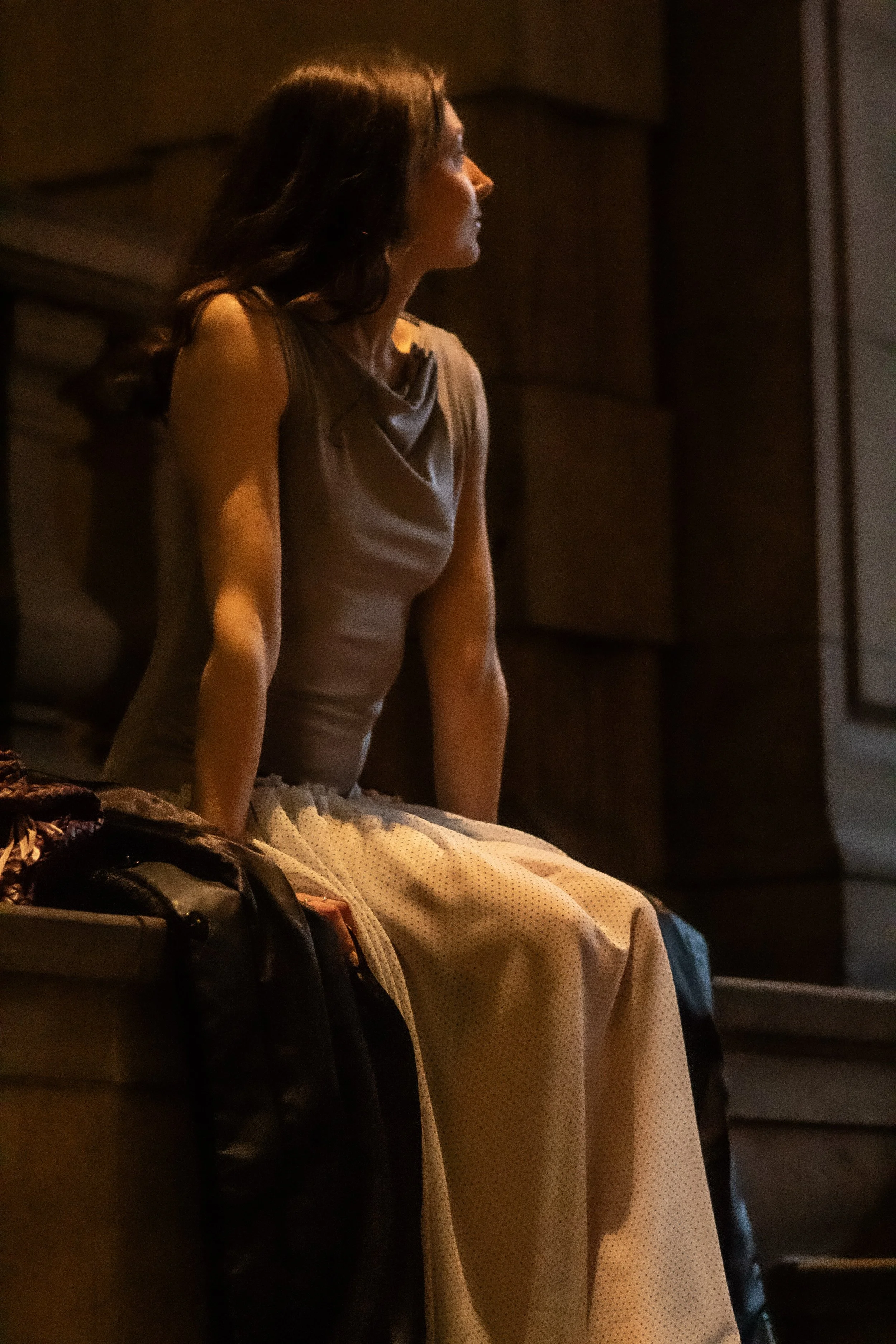 A woman with dark hair sitting on a bench in a dimly lit indoor space, wearing a sleeveless top and polka dot skirt.