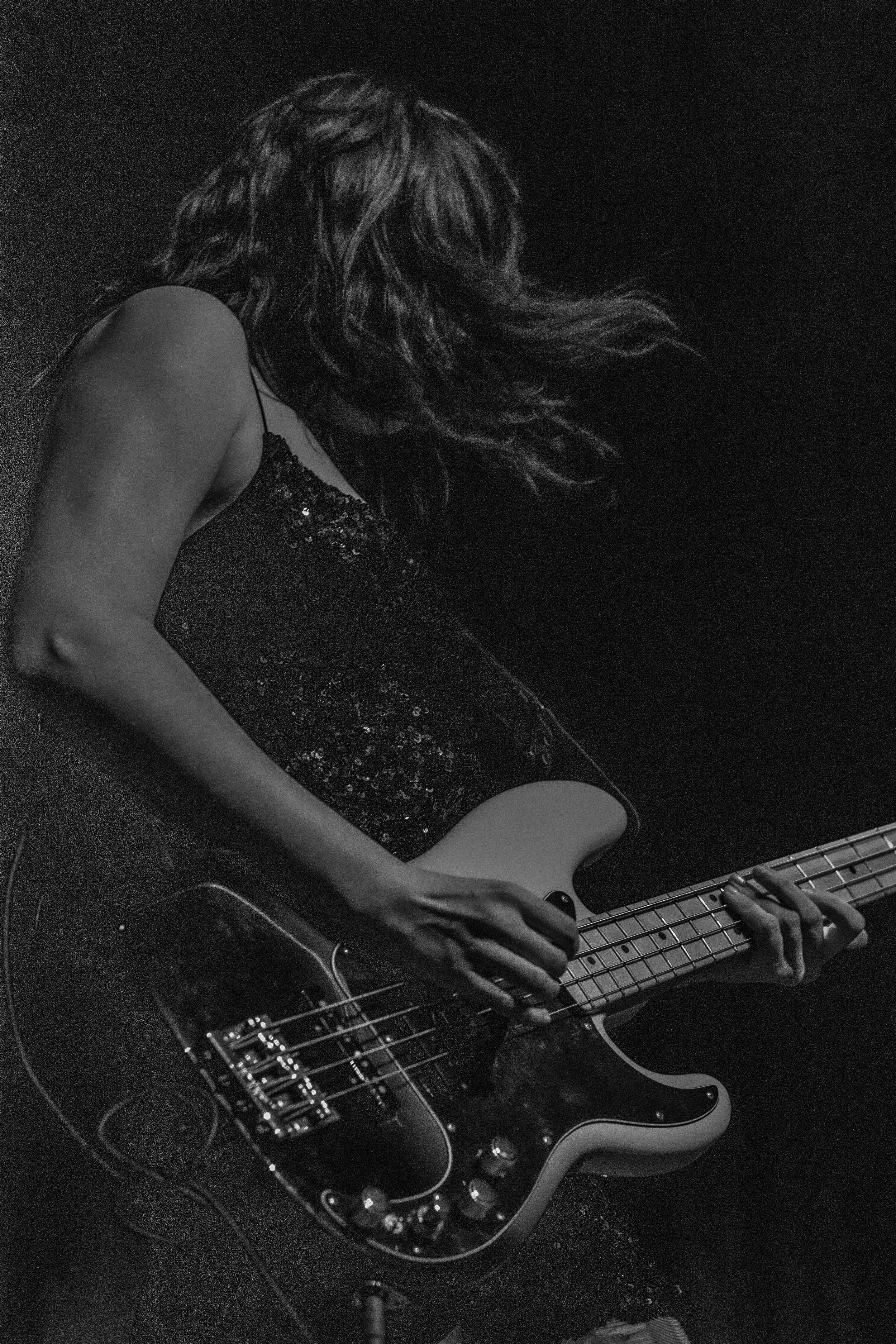 A woman playing an electric bass guitar in a dark setting, captured in black and white.