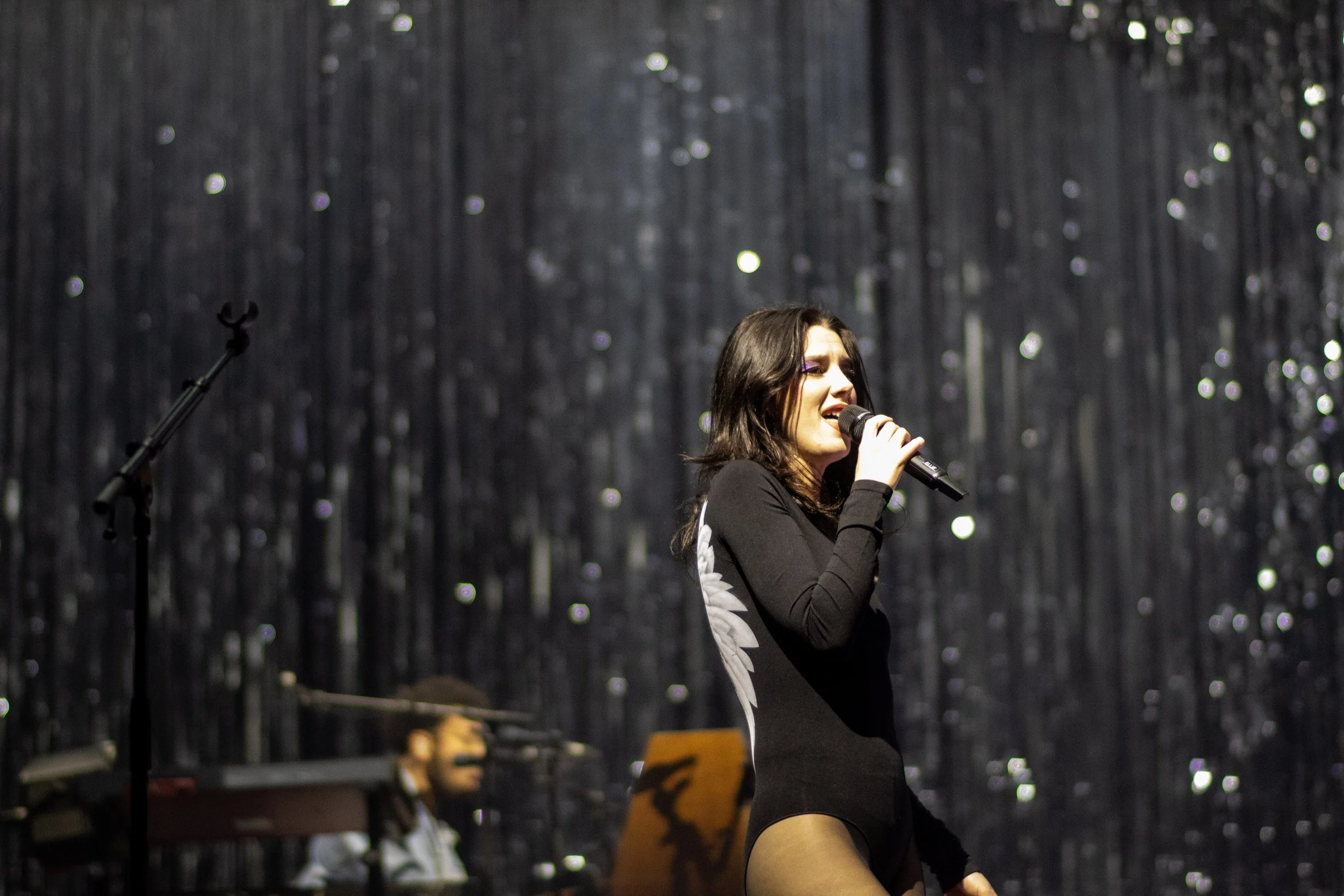 A female singer with dark hair performing on stage, holding a microphone, with a background of dark, shimmering curtains and a male musician playing keyboard in the background.