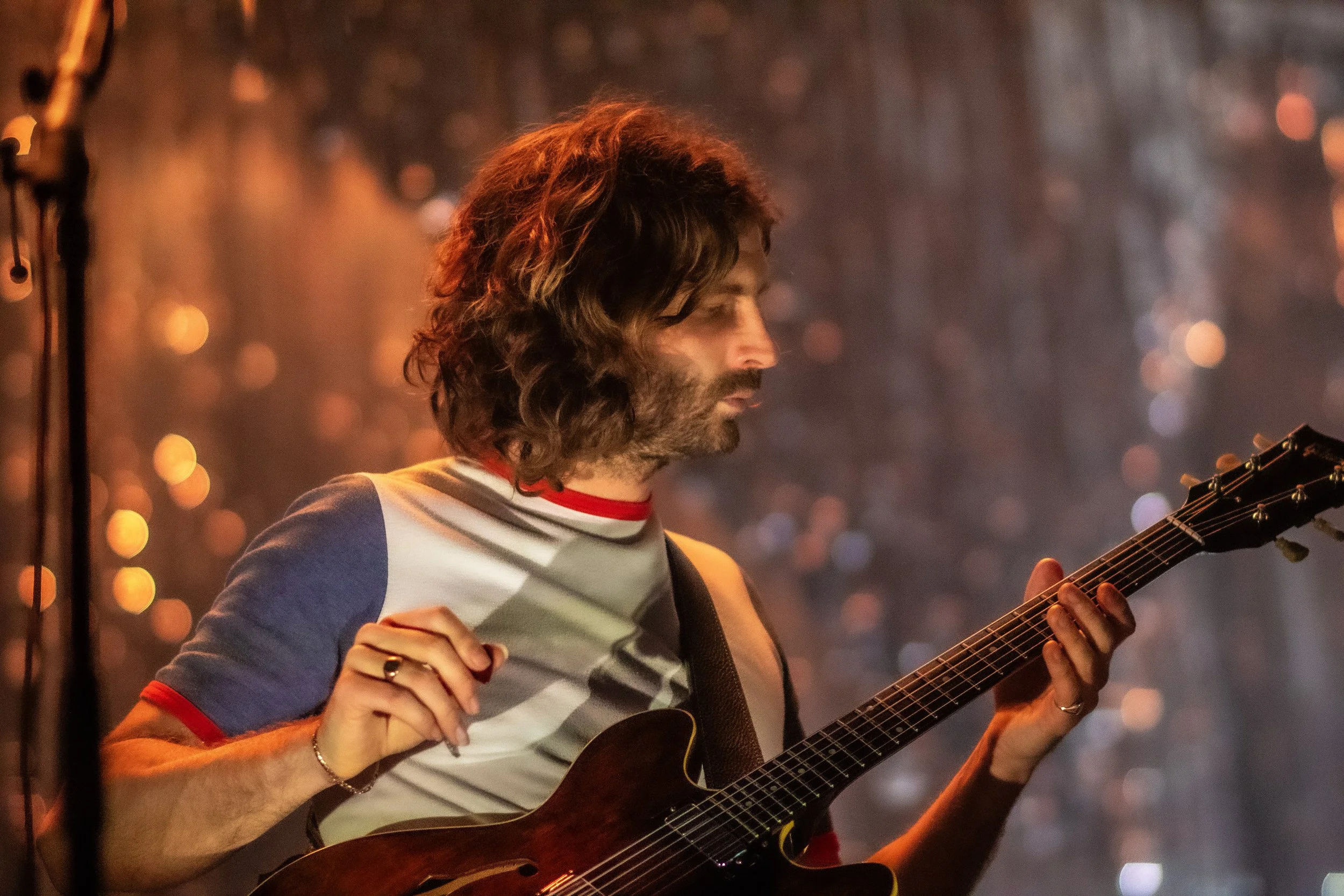 A man with long, wavy hair and a beard playing an electric guitar on stage, wearing a multicolored shirt, with a blurred background of stage lights.