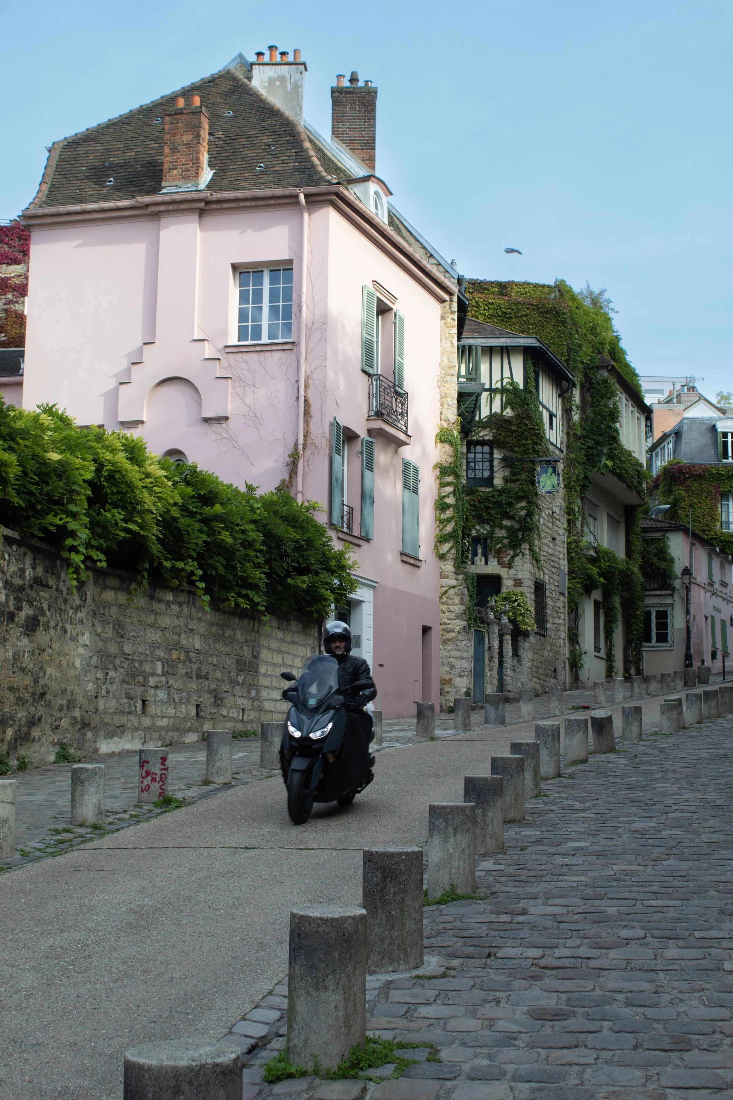A person riding a black scooter up a cobblestone street alongside a pink building with green shutters, with other buildings and greenery in the background.