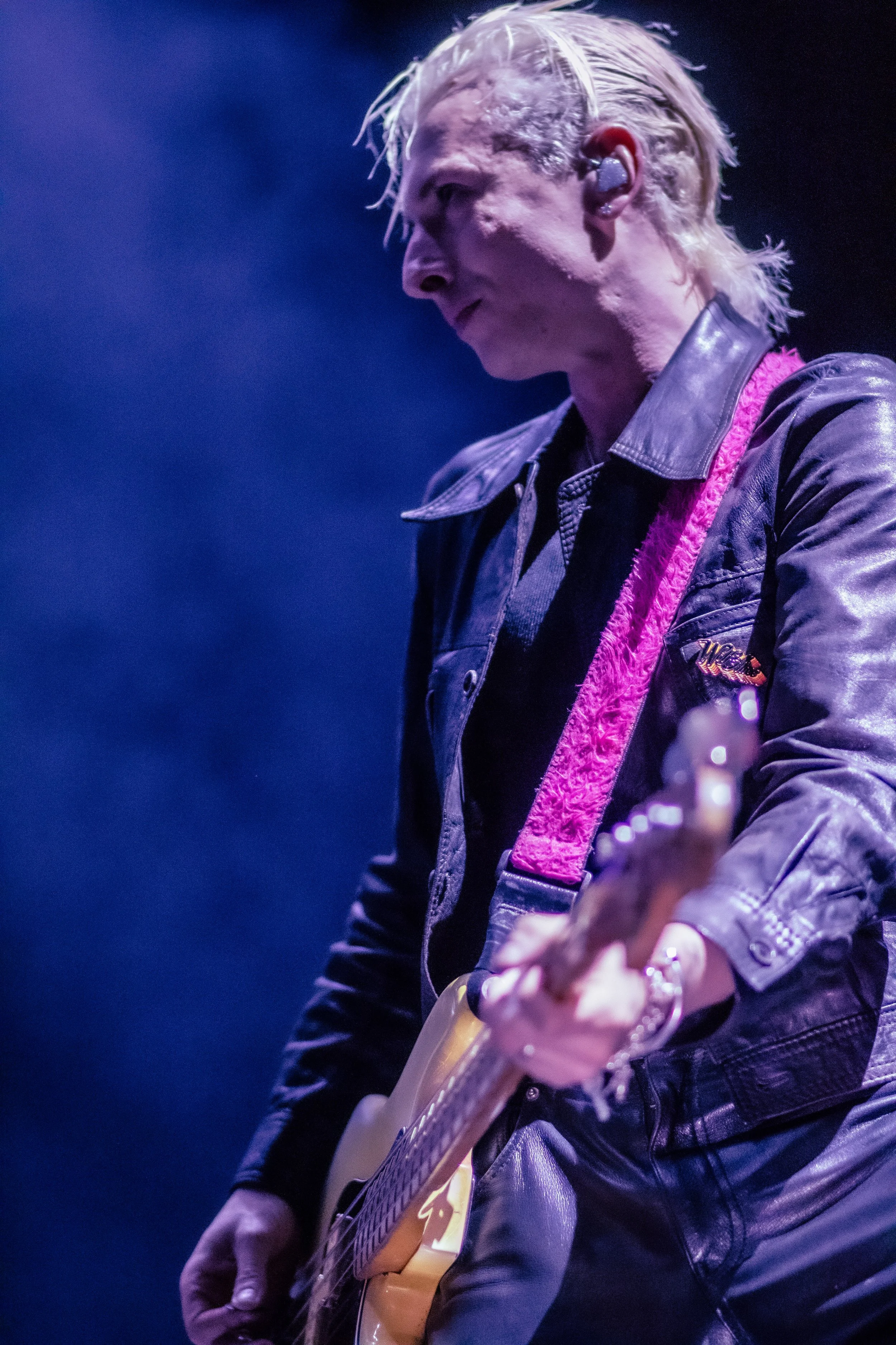 A musician playing an electric guitar on stage, wearing a black leather jacket with a pink strap, with blonde hair and in-ear monitors.