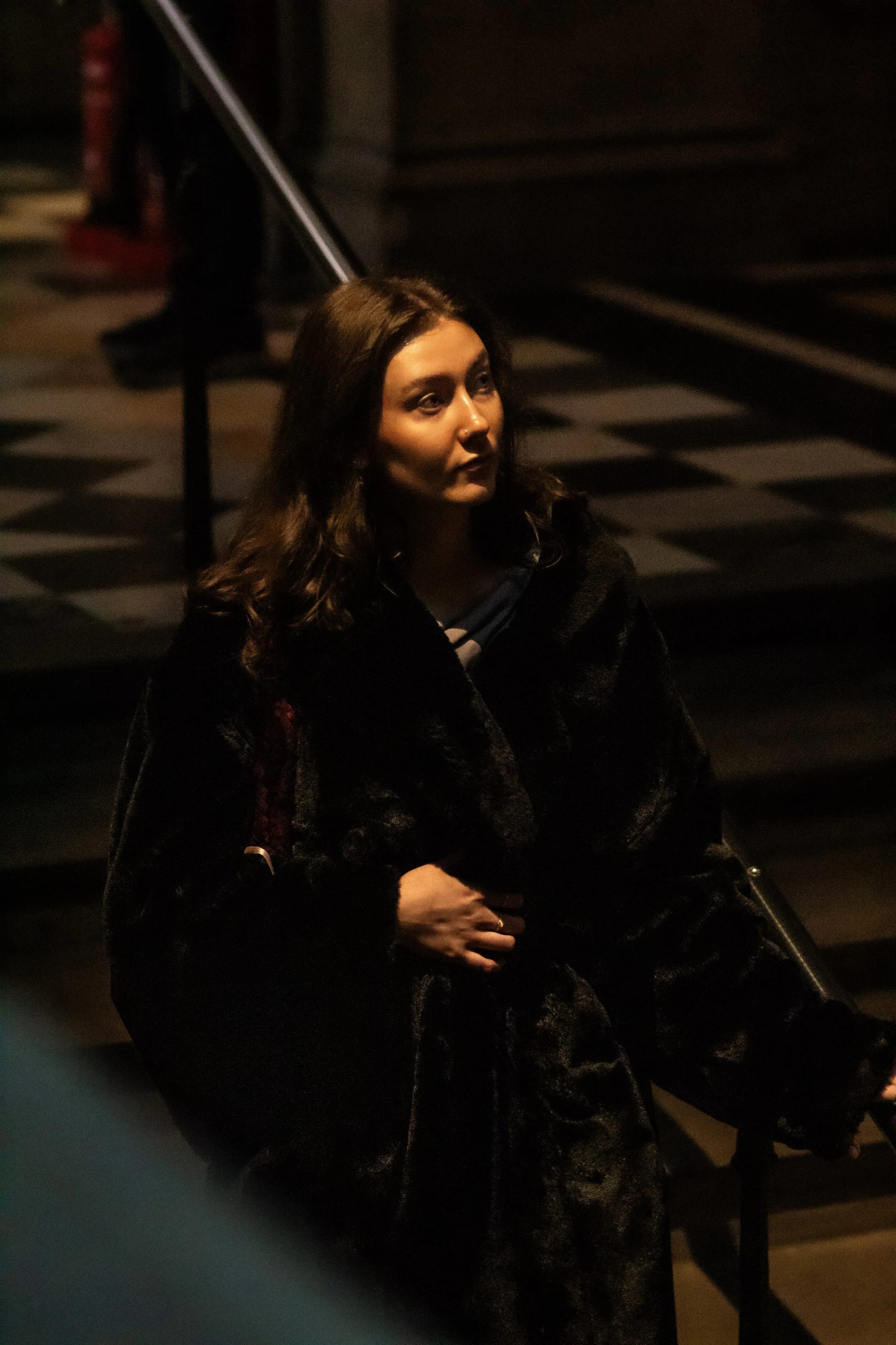 A woman sitting on stairs, illuminated by soft, warm lighting, wearing a dark coat, with a contemplative expression.