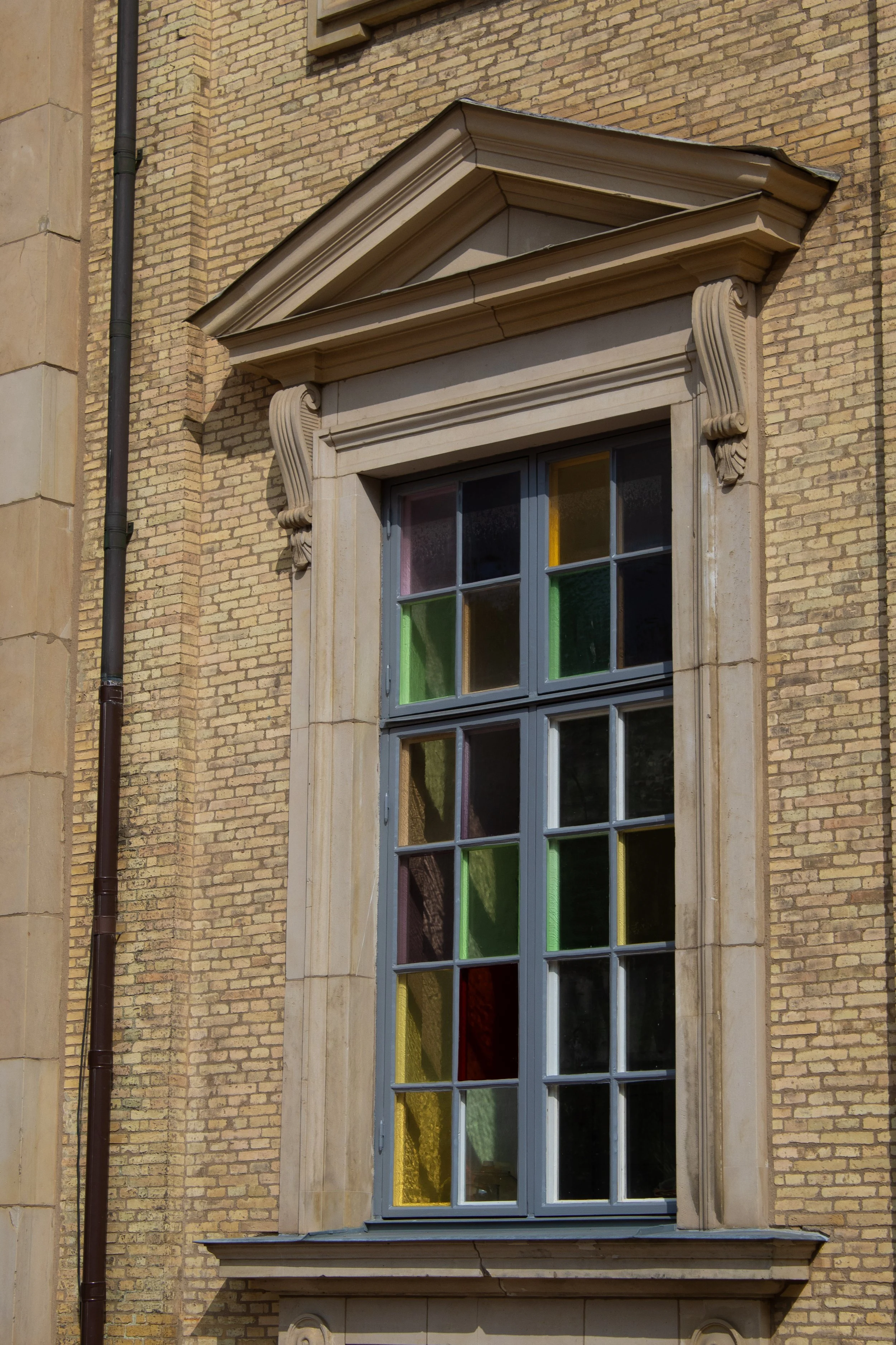 A brick building with a large window featuring multiple stained glass panels in various colors, framed by decorative stone trim and a small pediment on top.