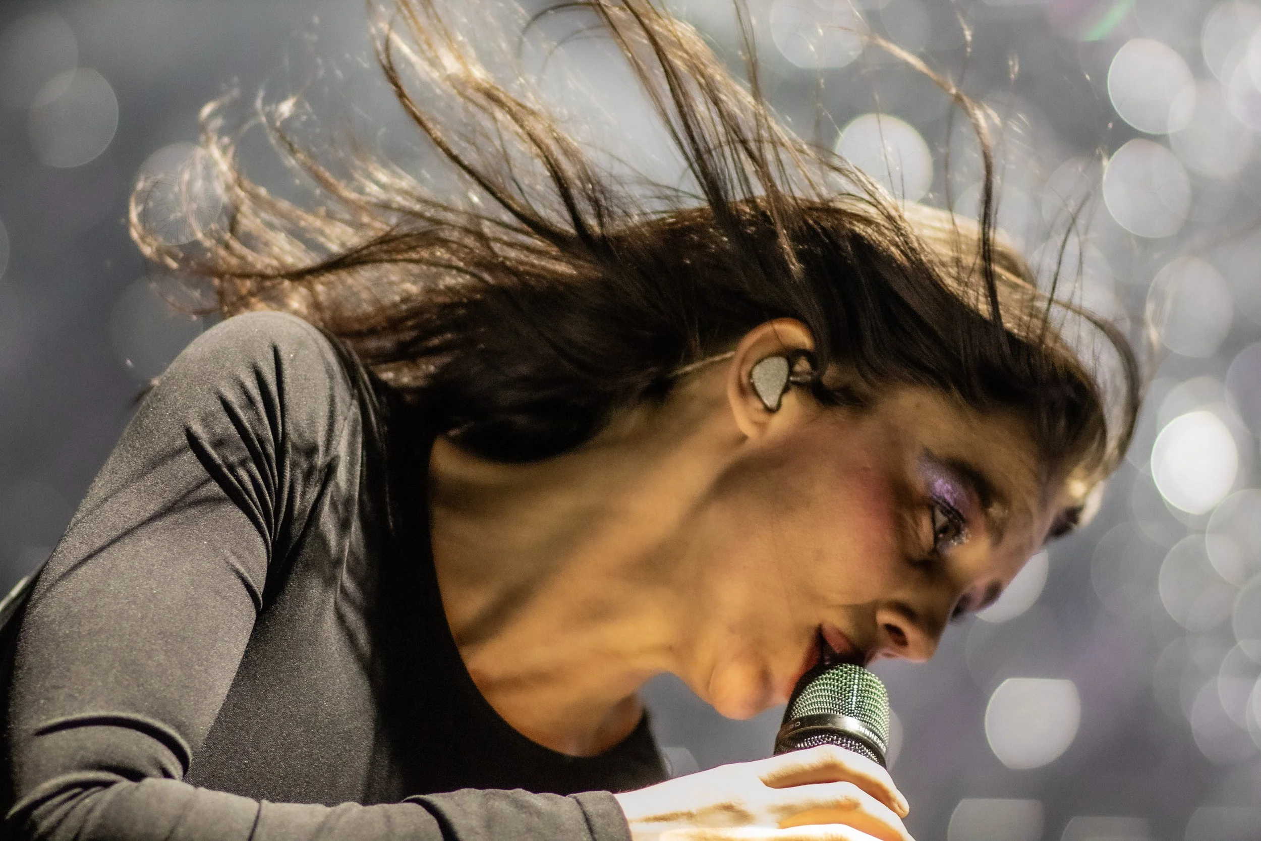 Close-up of a woman with long, dark hair singing into a microphone, wearing a in-ear monitor and a black long-sleeve top, with a blurred background of circular lights.