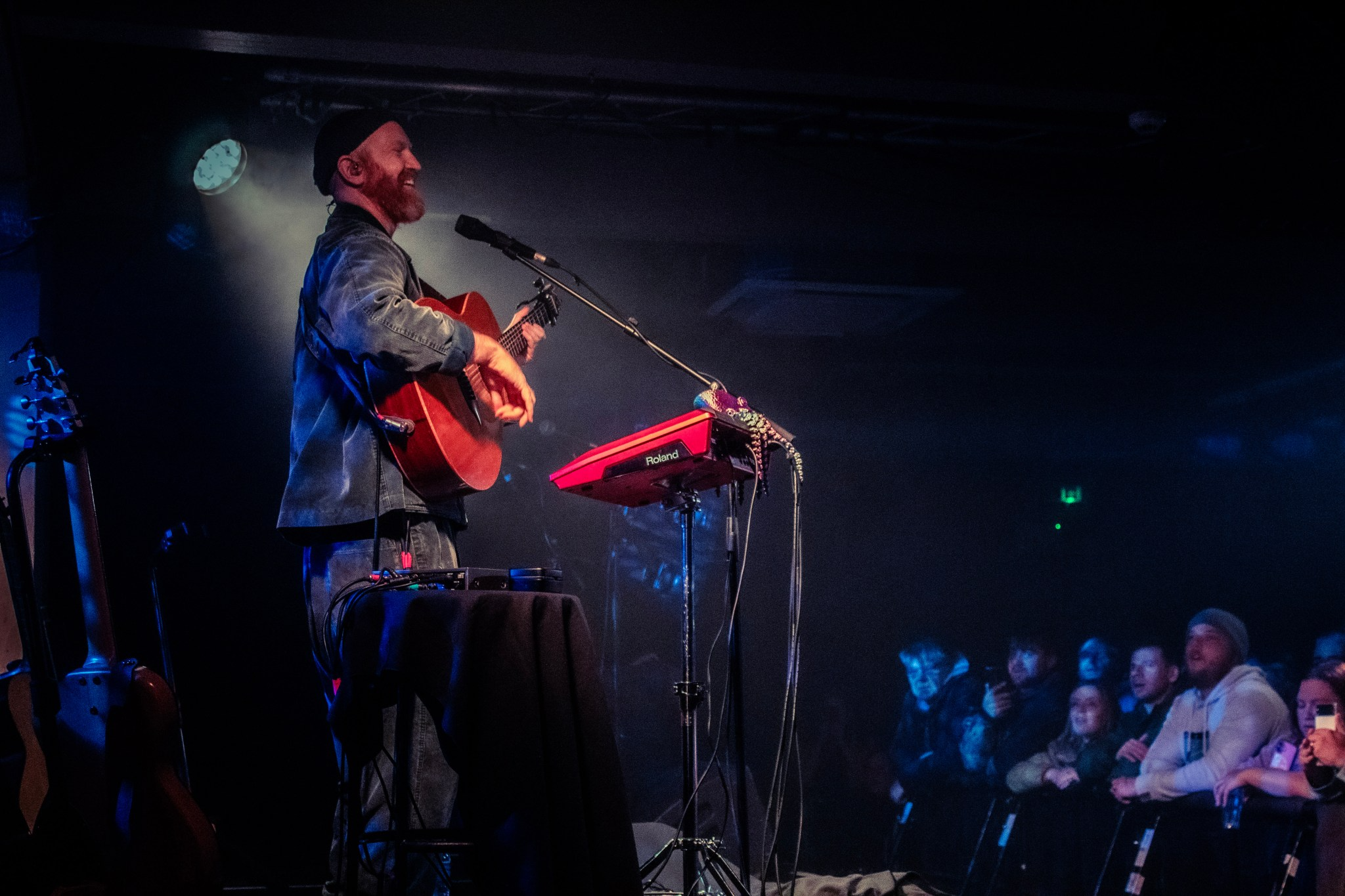 A musician with a beard playing an acoustic guitar on stage with a red keyboard in front of him, and an audience of people watching in the dark