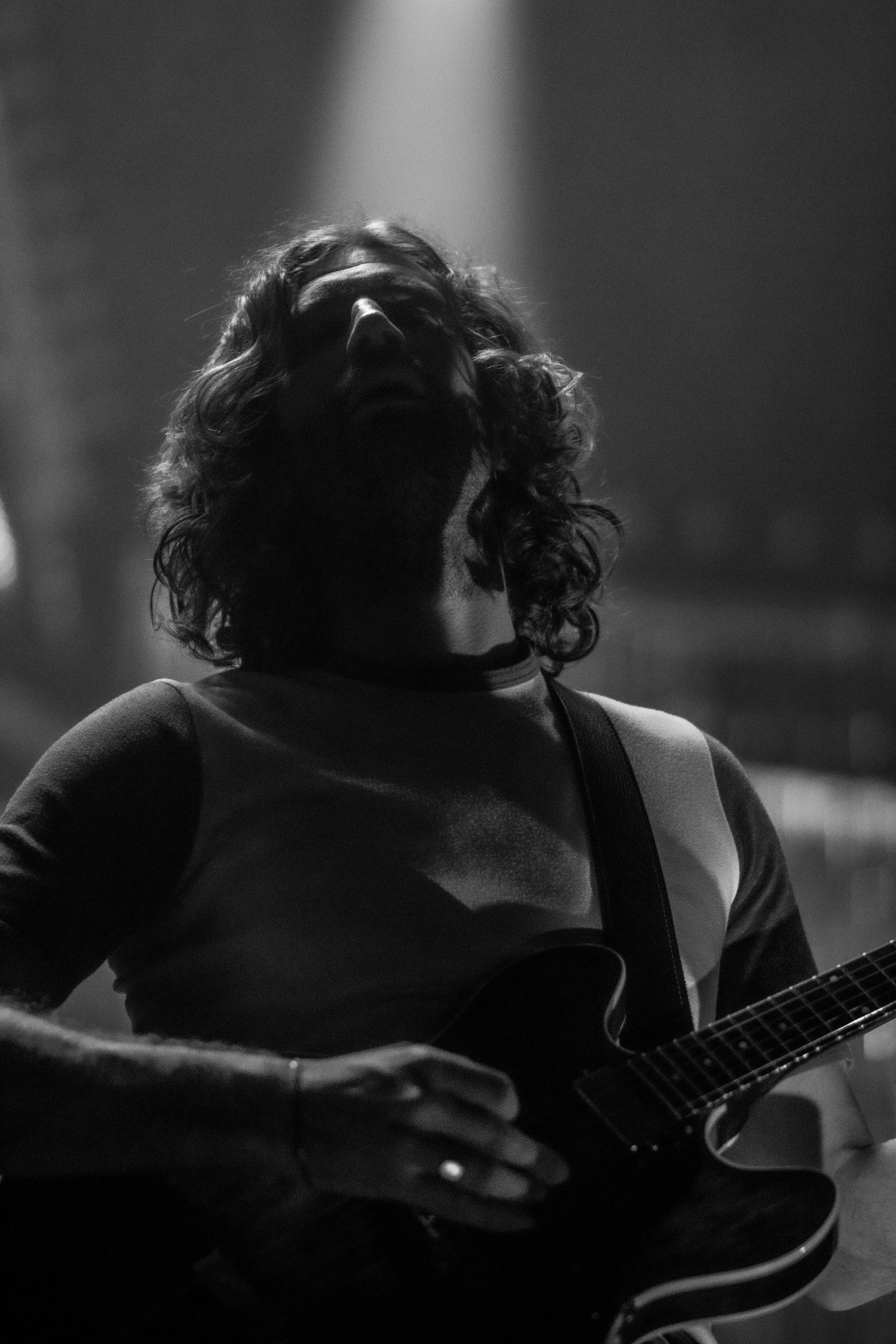 A man with long curly hair and glasses playing an electric guitar on stage under dramatic lighting.