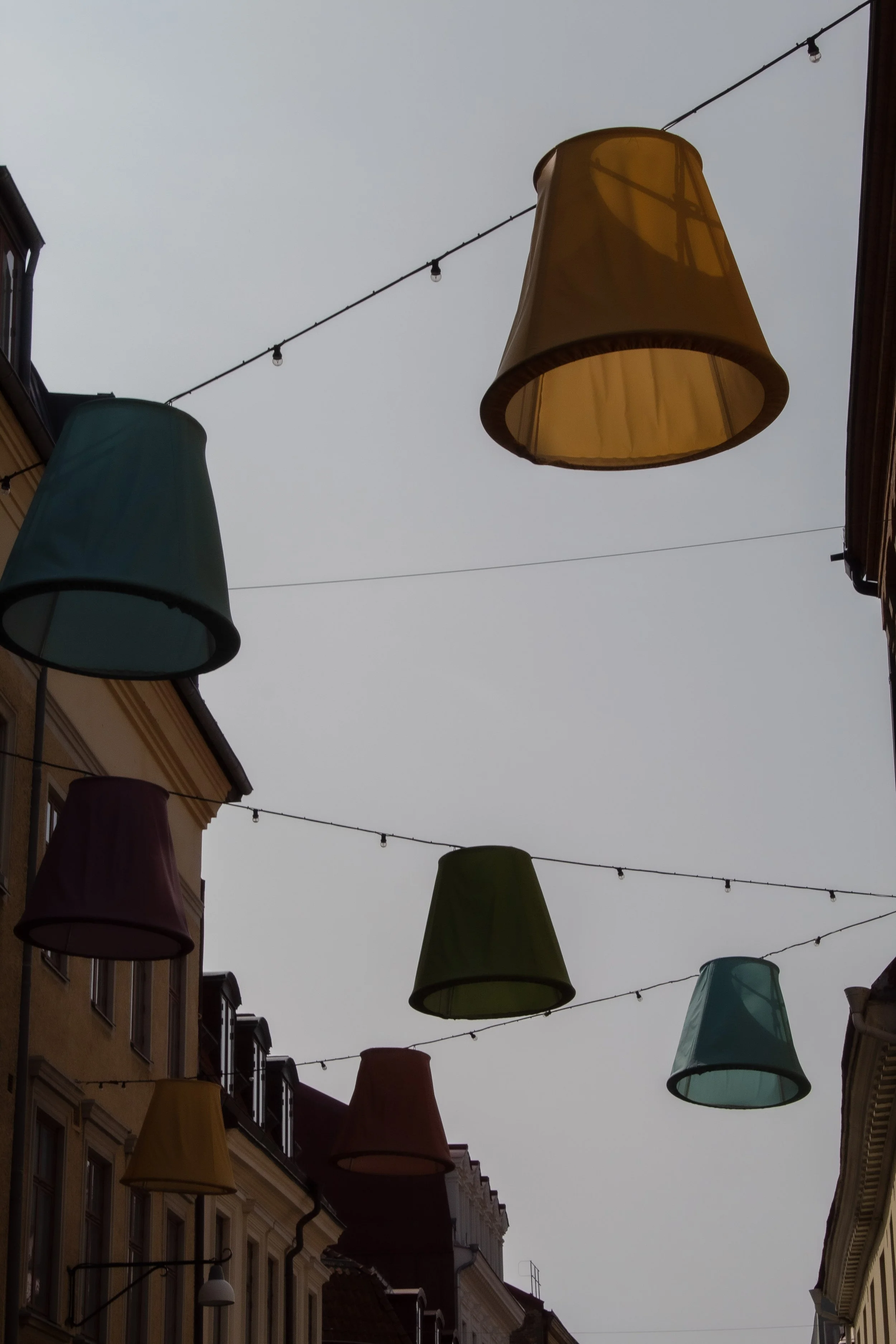Colorful lampshades hanging on strings across a city street with cloudy sky overhead.