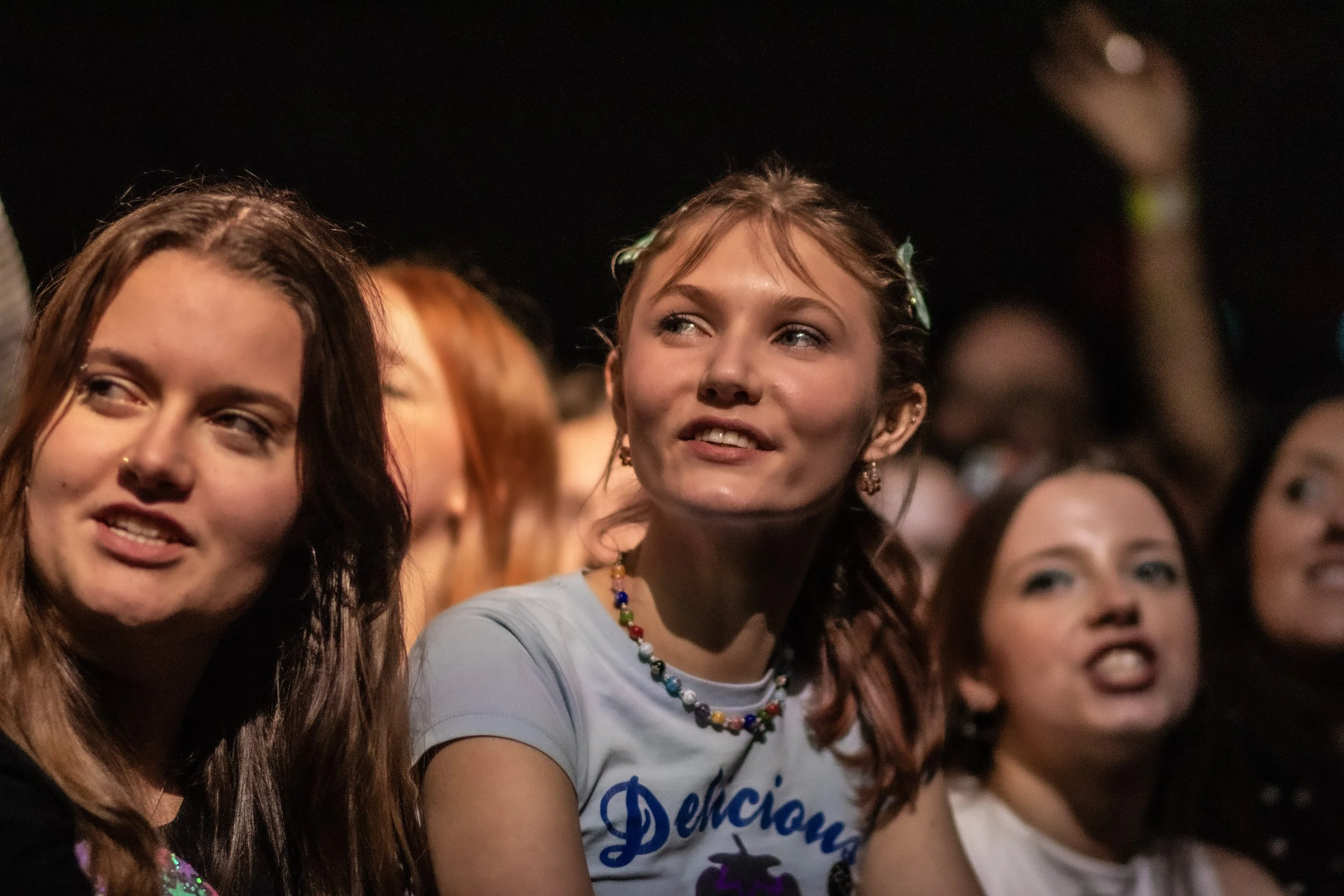 Group of young women at a concert or event, smiling and looking towards the stage. One woman wears a colorful beaded necklace and a shirt with text.